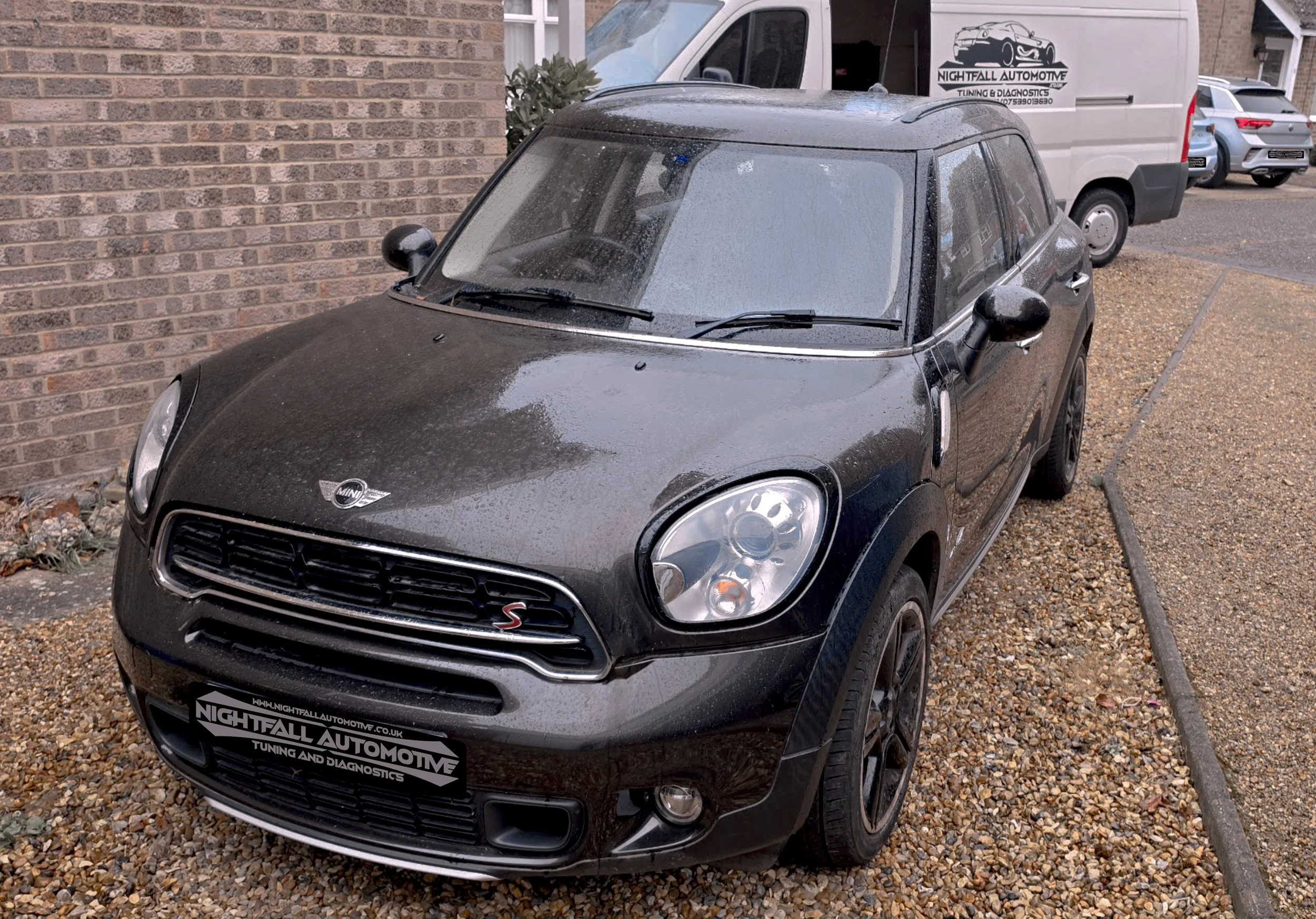 A black Mini Cooper S parked outside a building, with a wet windshield and a dusting of rain on the hood. A white van and other vehicles are visible in the background, along with a brick wall on the left.