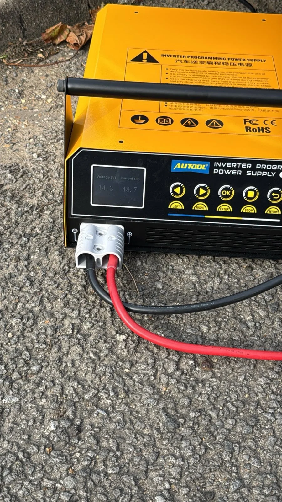 A yellow and black inverter power supply device with a digital display showing voltage and current, connected to red and black cables on a paved surface.