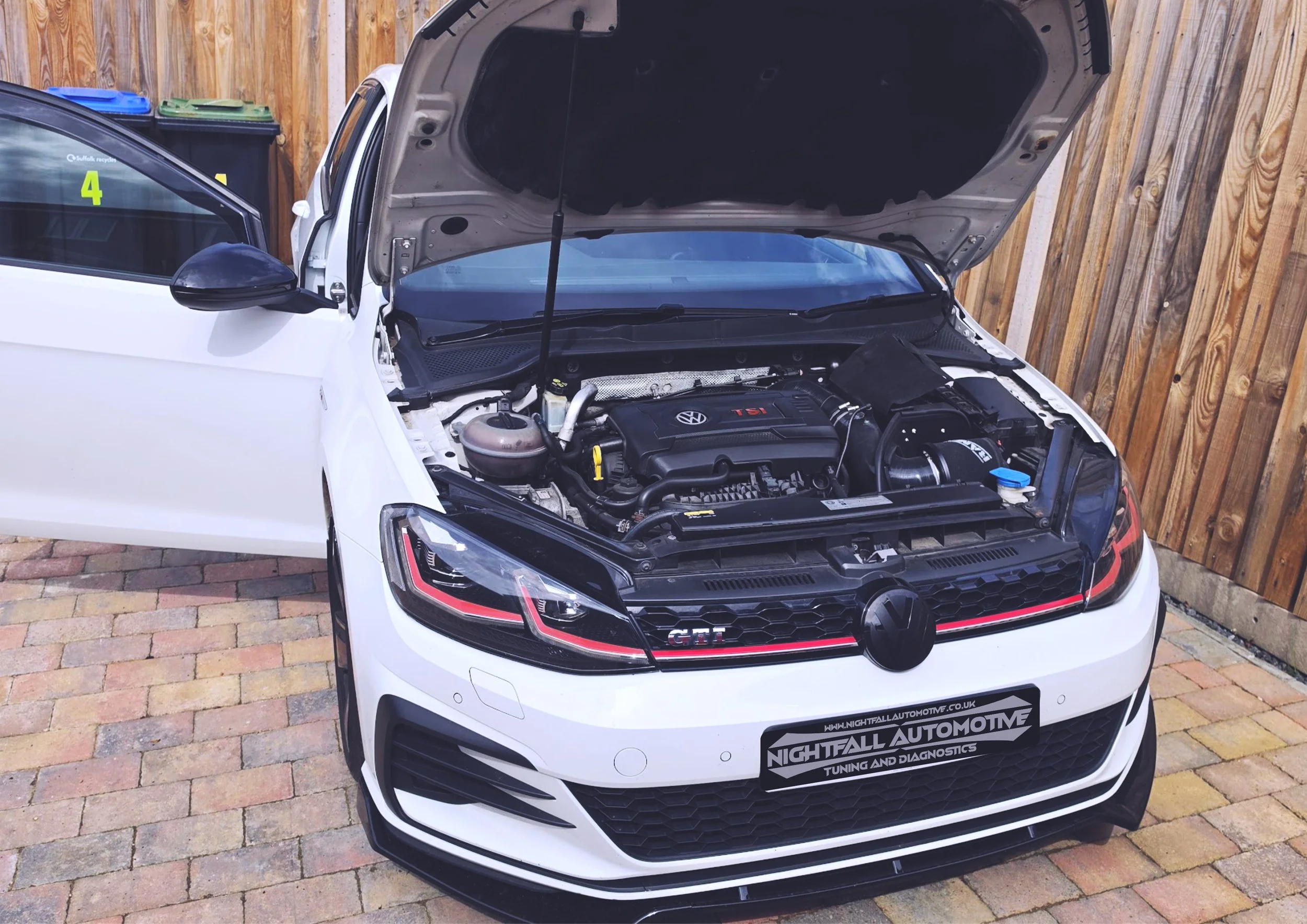 A white Volkswagen car with its hood open, parked on a paver driveway next to a wooden fence. The car has a North Face USA sticker on the windshield, and a sign on the front bumper reads "Nightfall Automotive Tuning and Diagnostics".