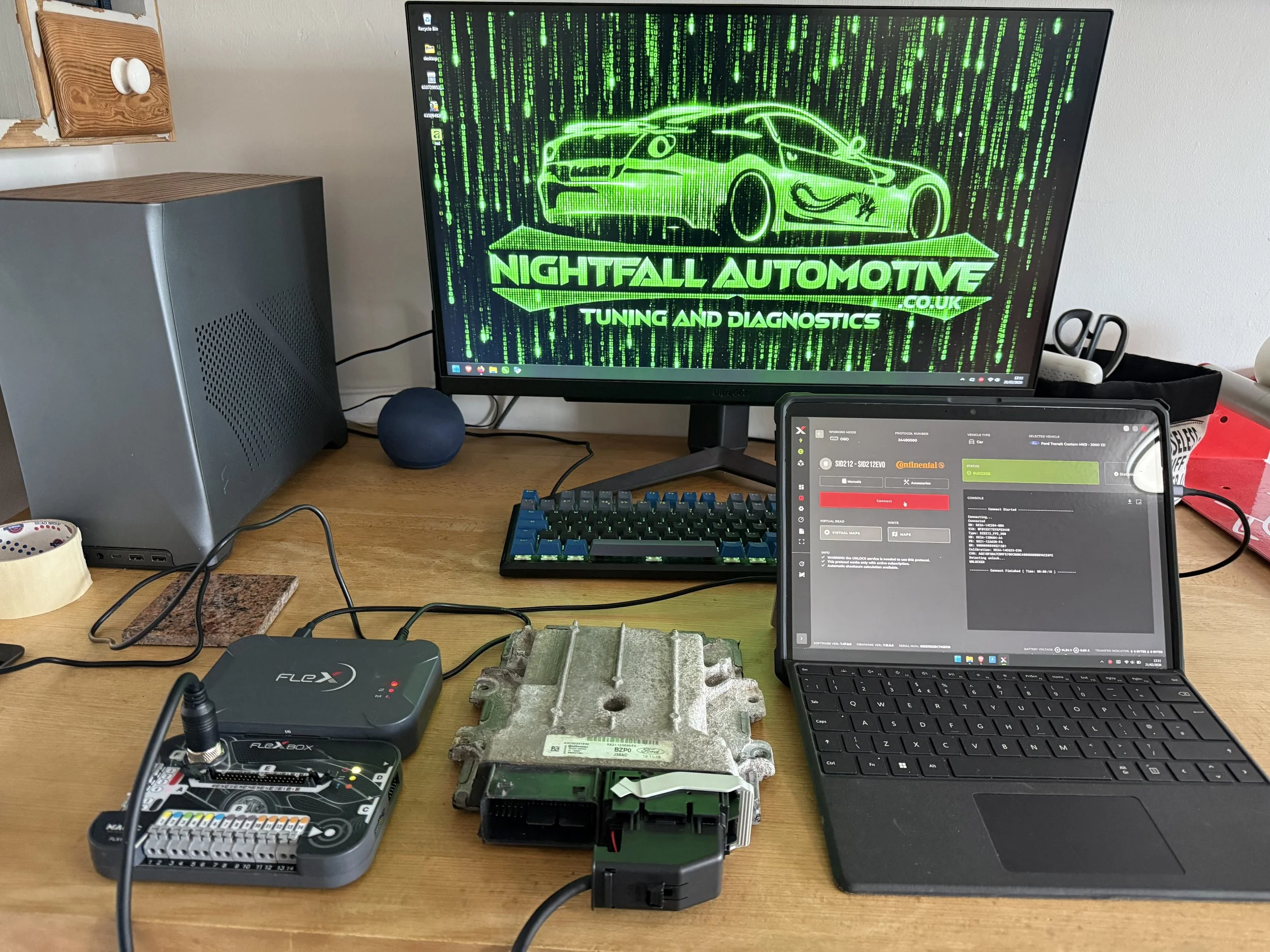 A workspace with a computer monitor displaying a green digital car outline and text related to automotive tuning, and an open laptop showing diagnostic software. The desk also has electronic devices, cables, a small speaker, and a metal engine contro