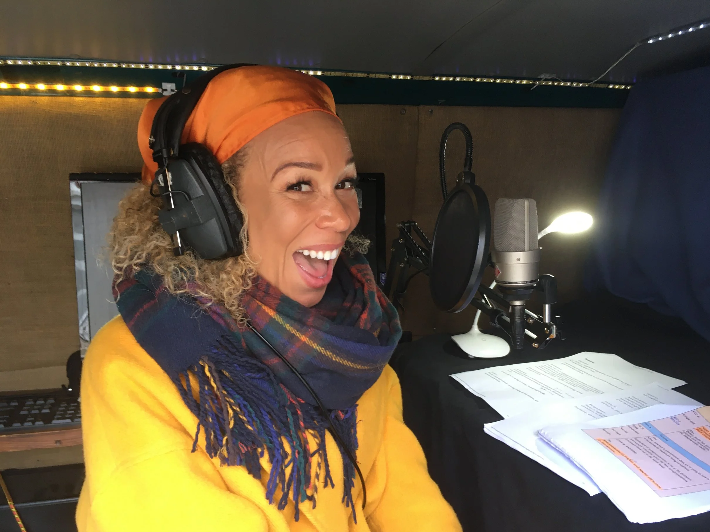 A woman with curly blond hair, wearing a bright yellow sweater, a multicolored scarf, and an orange headscarf, is smiling widely in a  mobile recording van with a microphone, headphones, and papers on a desk in front of her.