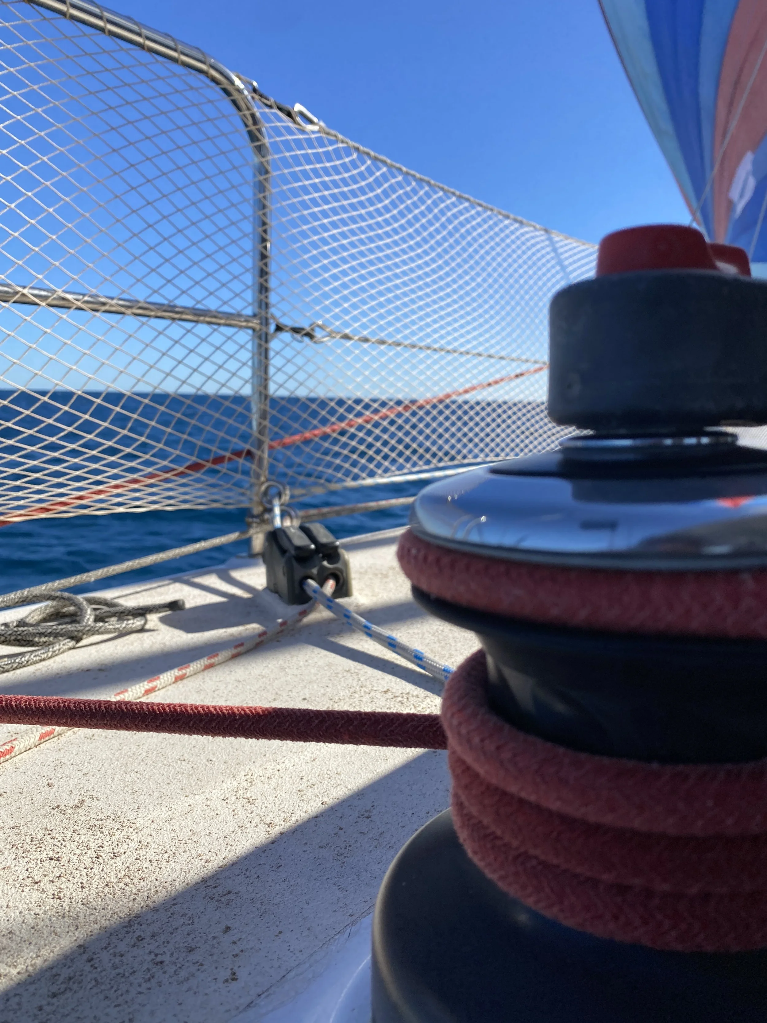 Close-up of a winch with red and black ropes on a sailboat, with a metal safety net and the ocean in the background under a clear blue sky.