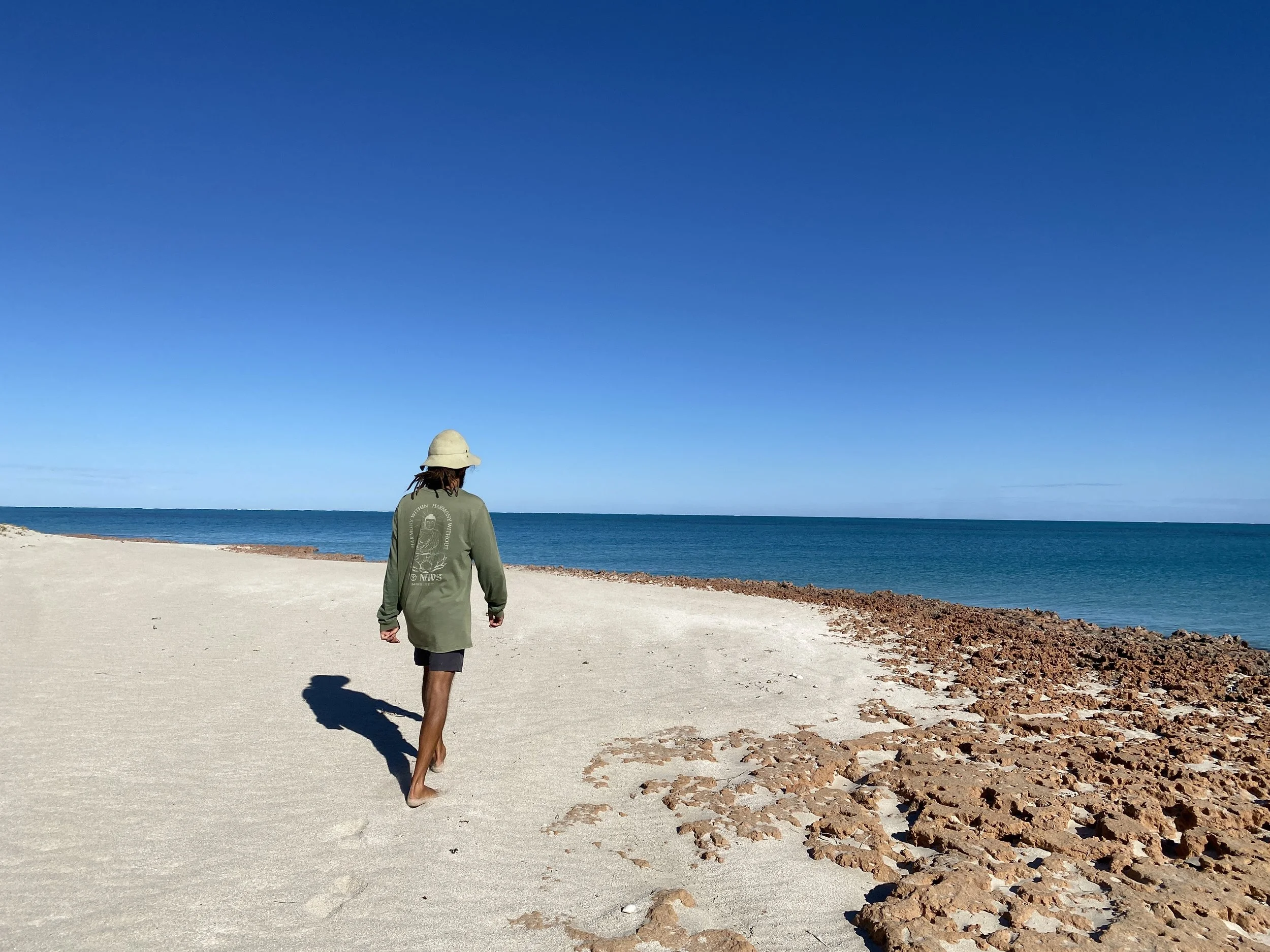Person walking alone on a sandy beach with rocks, under a clear blue sky, near calm ocean waters.
