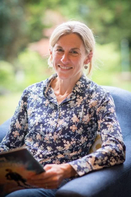 Philippa Hawkins is sitting in a comfortable chair looking at the camera holding a book