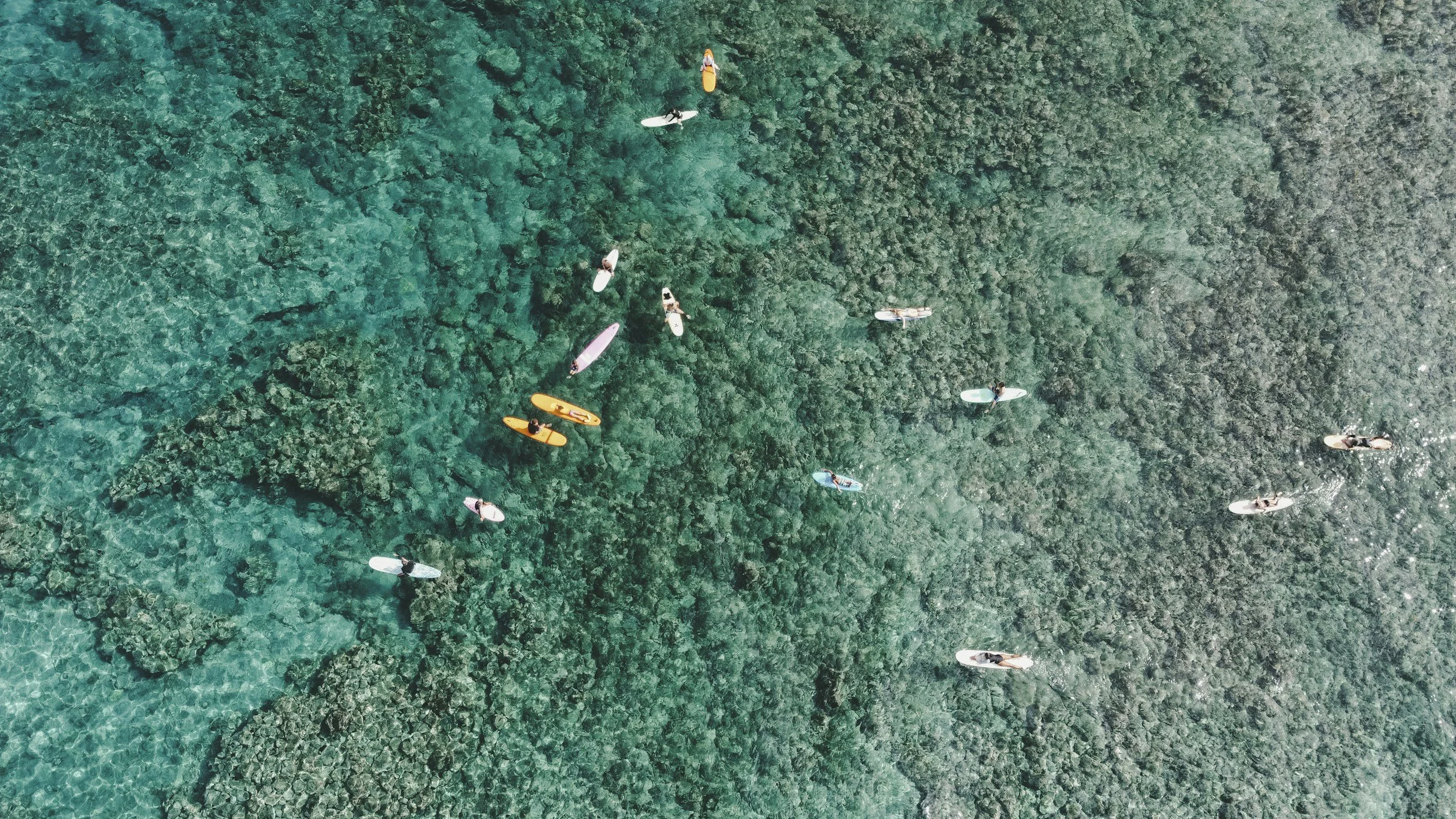Aerial view of multiple people surfing and paddleboarding on clear turquoise ocean water over a rocky reef.