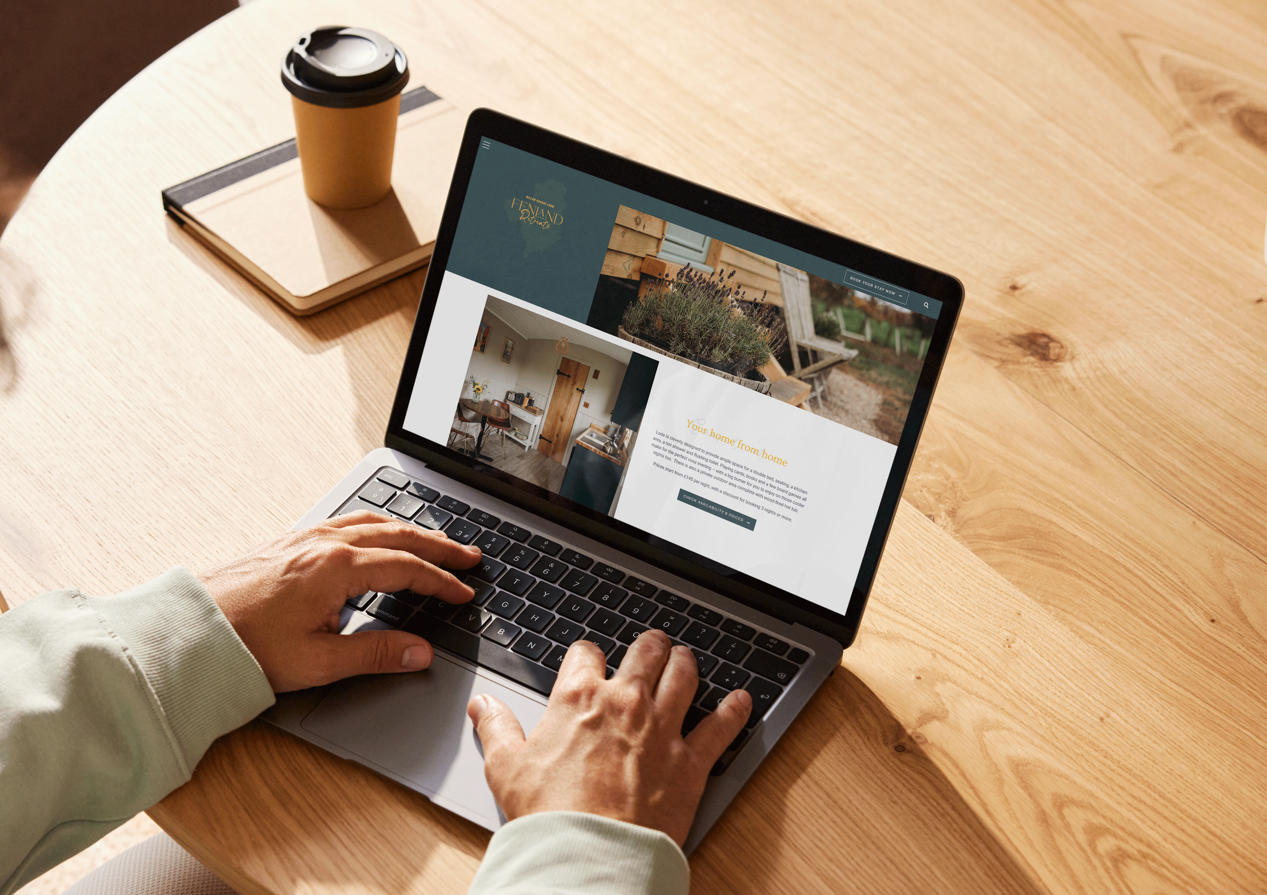 Person using a laptop on a wooden table with a cup of coffee, a notebook, and a folder nearby, browsing a glamping website.