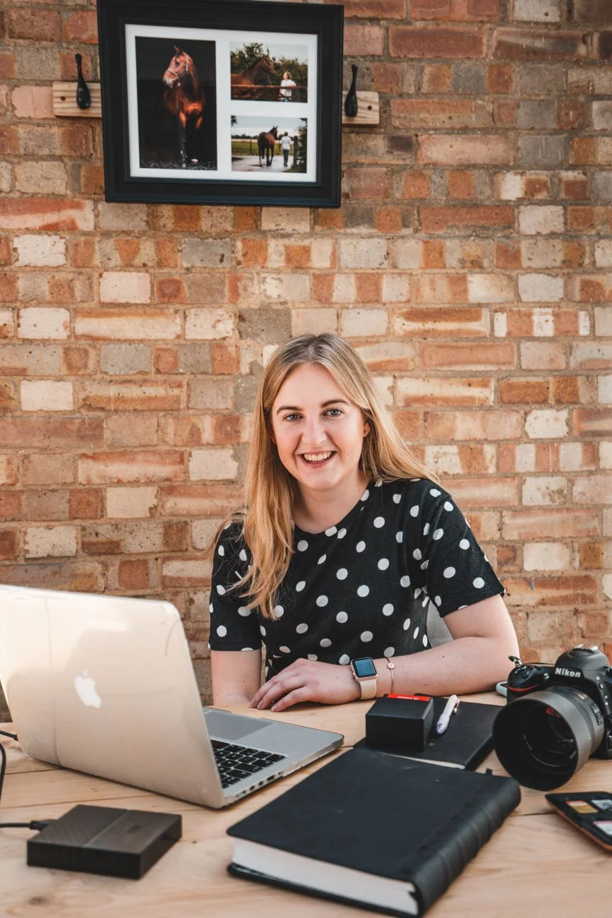 A young woman with long blonde hair sitting at a wooden desk with a silver MacBook, black notebook, camera, and other items. She is smiling and wearing a black polka dot shirt. Behind her is a brick wall with a framed collage of outdoor photos of horses.