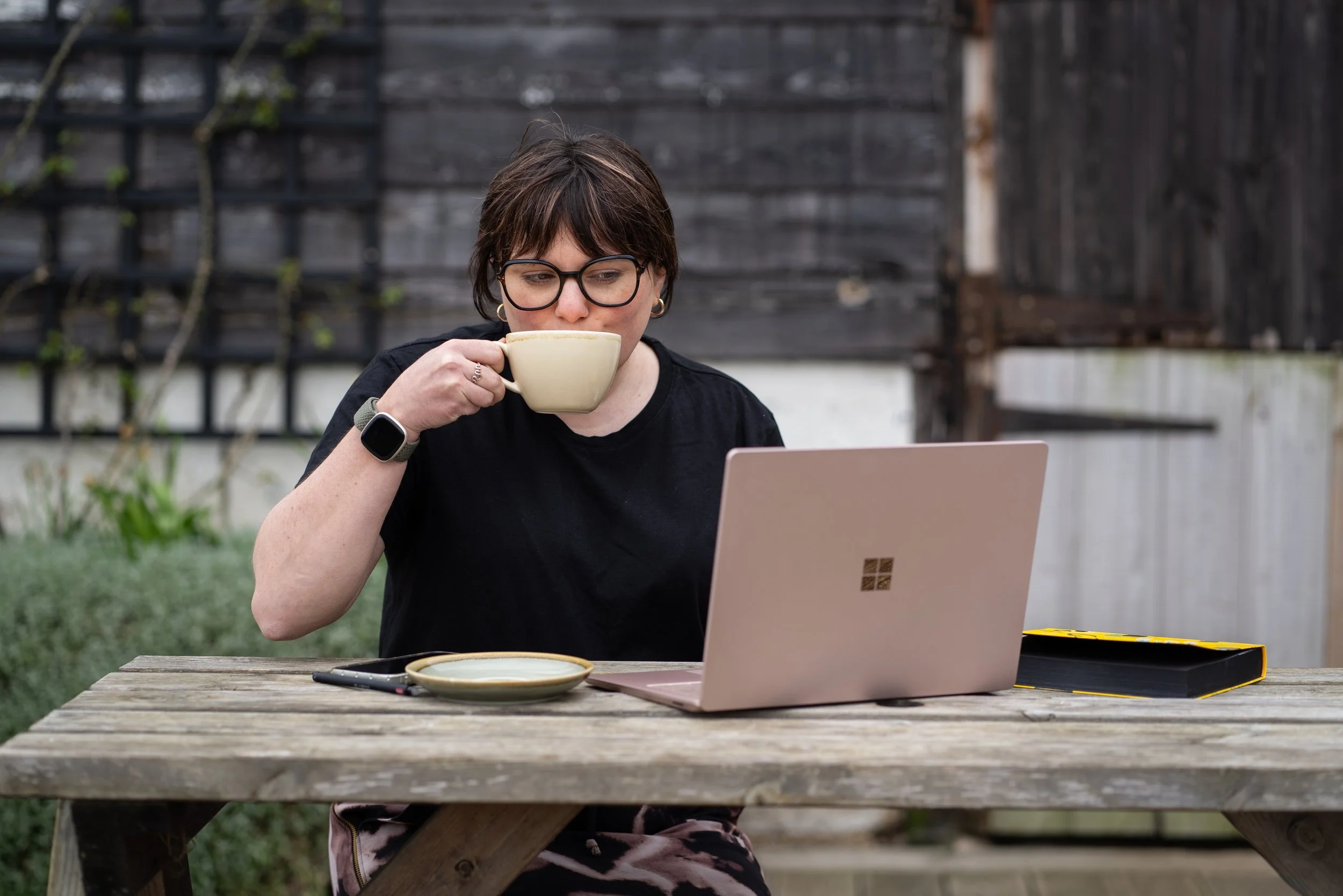 Virtual Assistant in black shirt sitting at outdoor wooden table with laptop, mug, and notebook, drinking coffee.