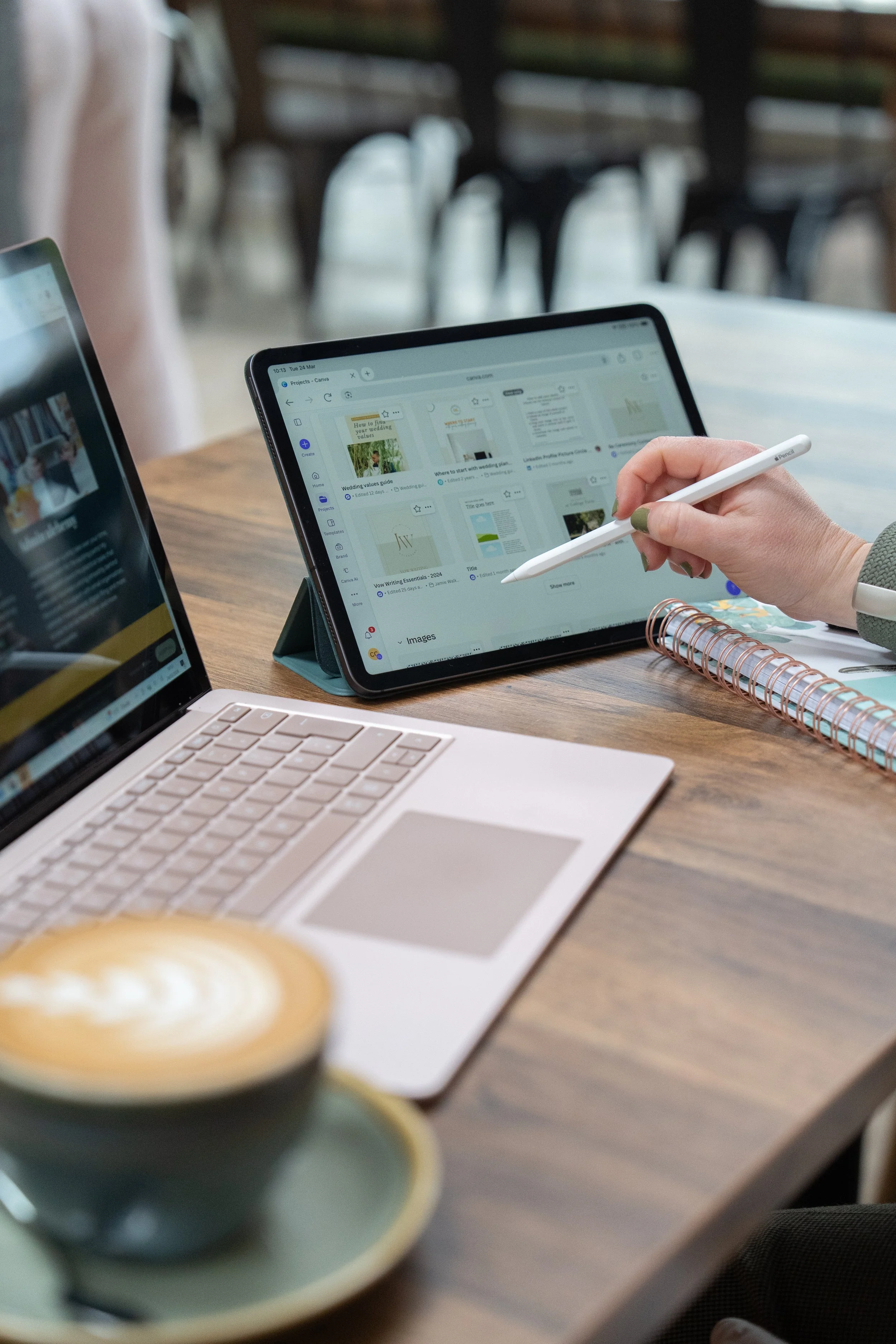 Person working on a tablet at a wooden table with a laptop and a cup of coffee nearby.