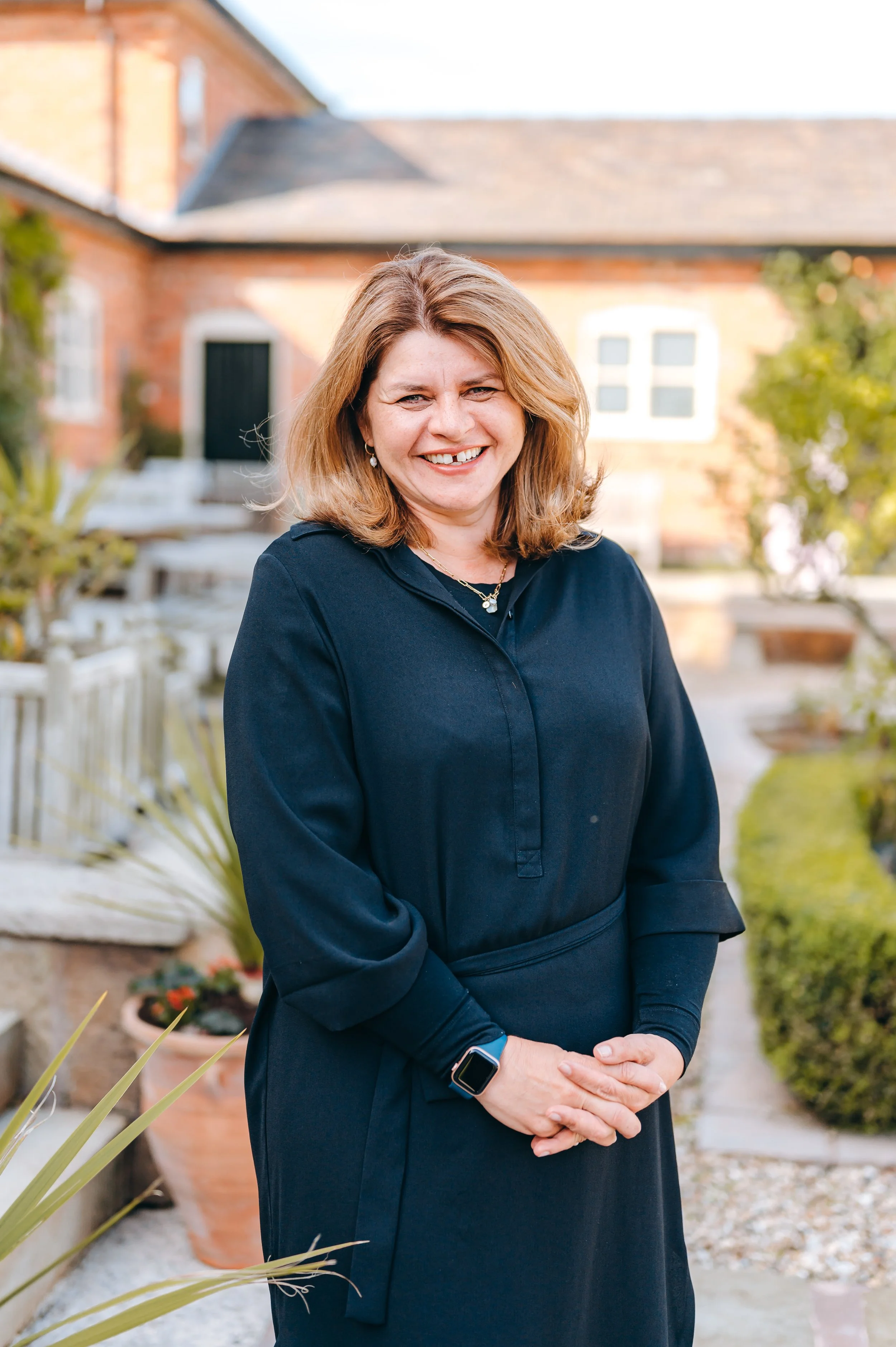 A woman with shoulder-length blond hair, wearing a navy blue long-sleeve top, standing outdoors in a garden with plants and a brick house in the background, smiling at the camera.