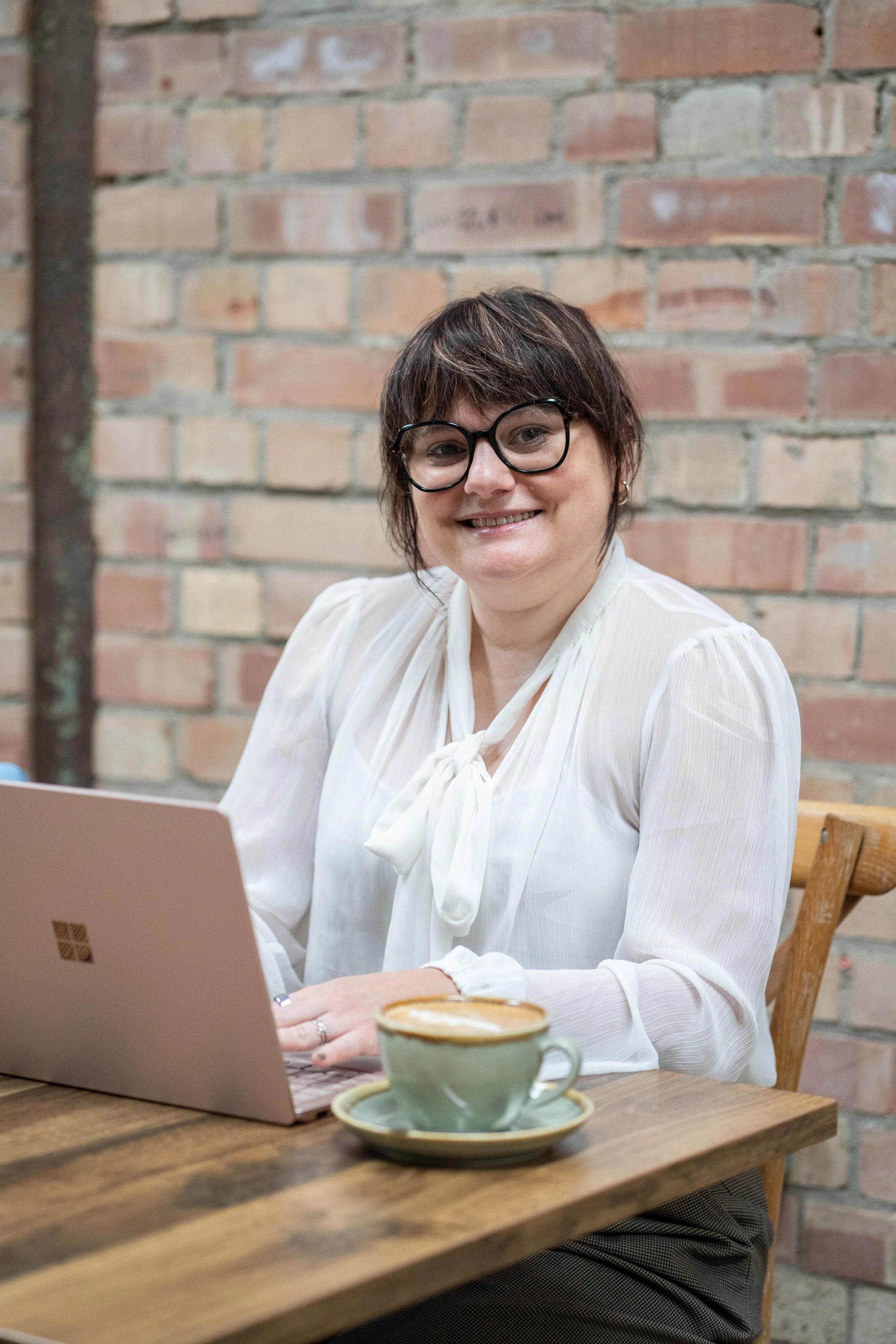 A white woman with short brown hair and glasses, wearing a white blouse, sitting at a wooden table, smiling at the camera, with a laptop in front of her and a cup of coffee on a saucer.