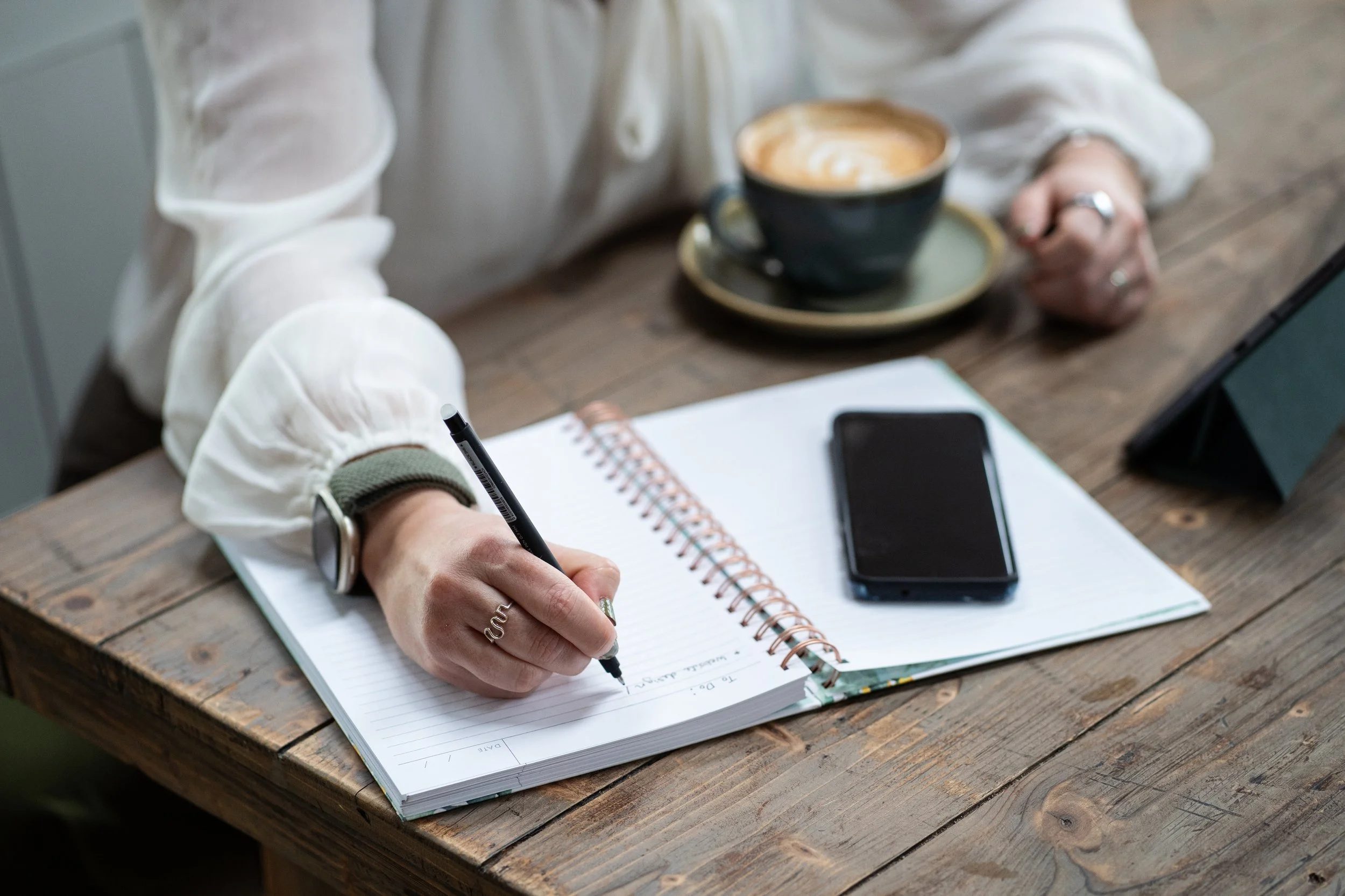 Virtual Assistant writing in a spiral notebook with a black pen, a smartphone, and a tablet on a rustic wooden table. A cup of coffee in a black mug on a saucer is also on the table.