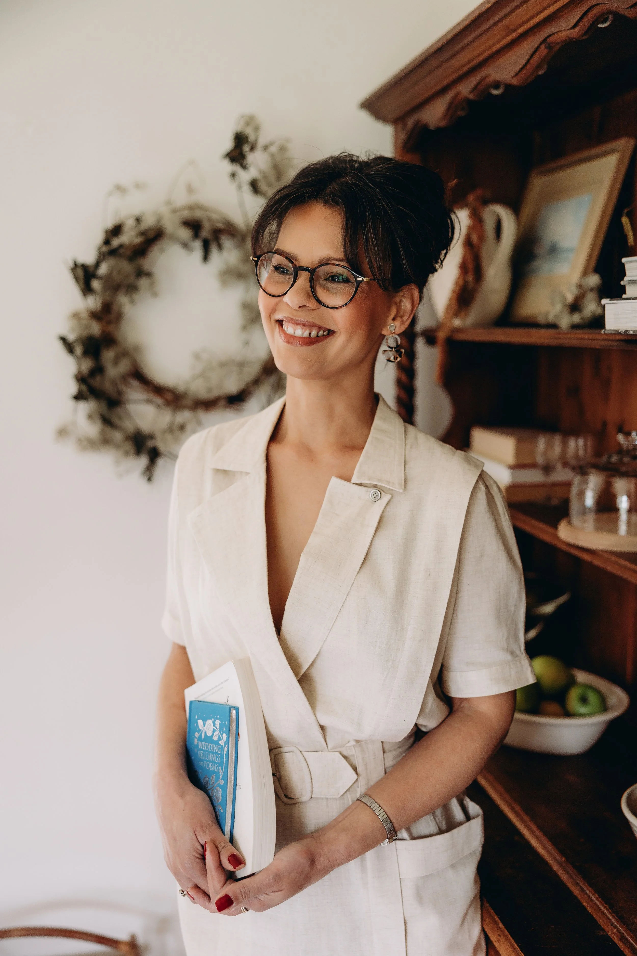 A smiling woman with short dark hair, glasses, and earrings stands indoors, holding a book and some papers, with a wooden shelf and a decorative wreath in the background.