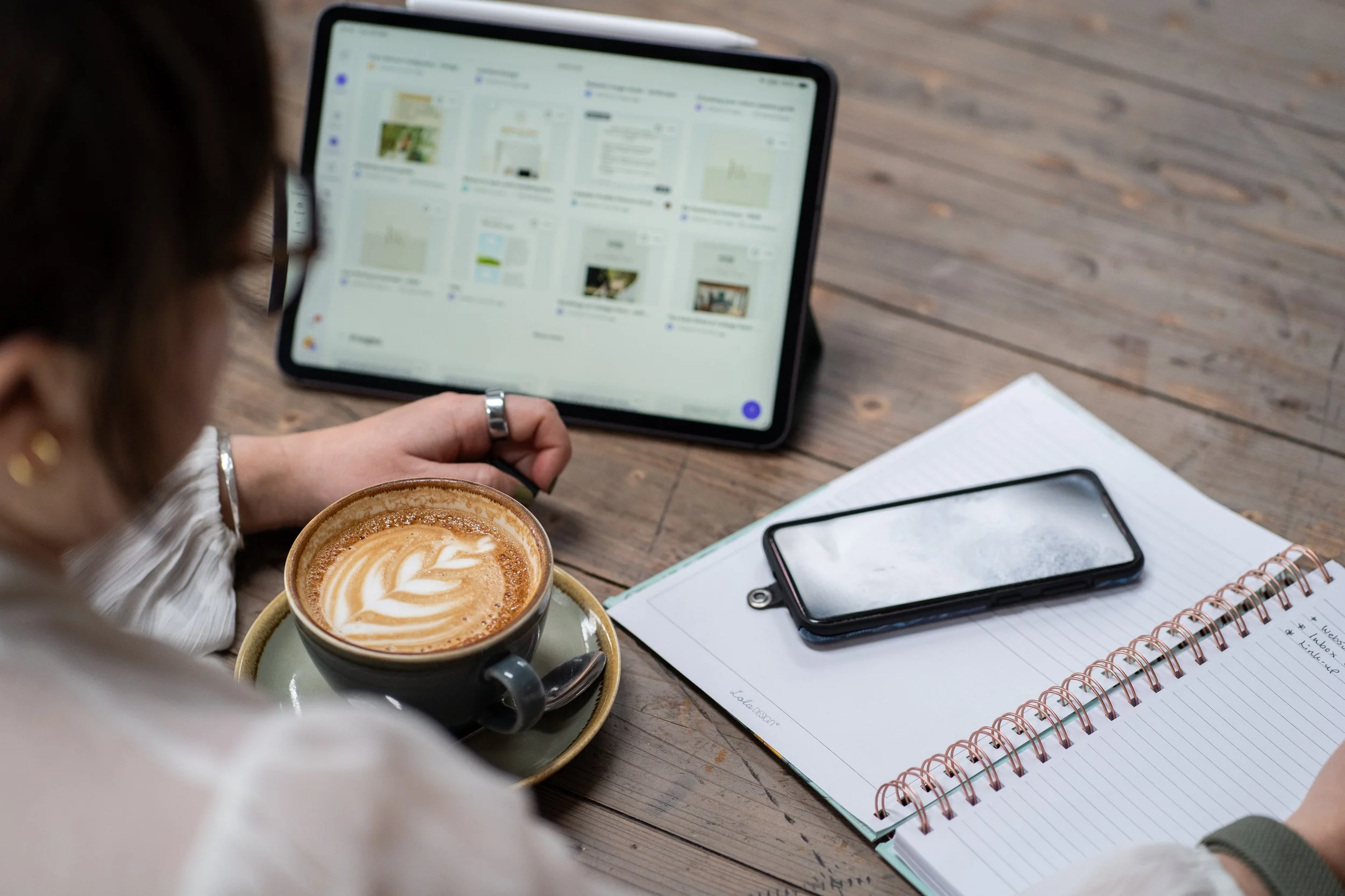 Virtual Assistant sitting at a wooden table with a cup of coffee, a smartphone, a tablet, and a notebook. The person is looking at the tablet screen while holding the coffee cup.