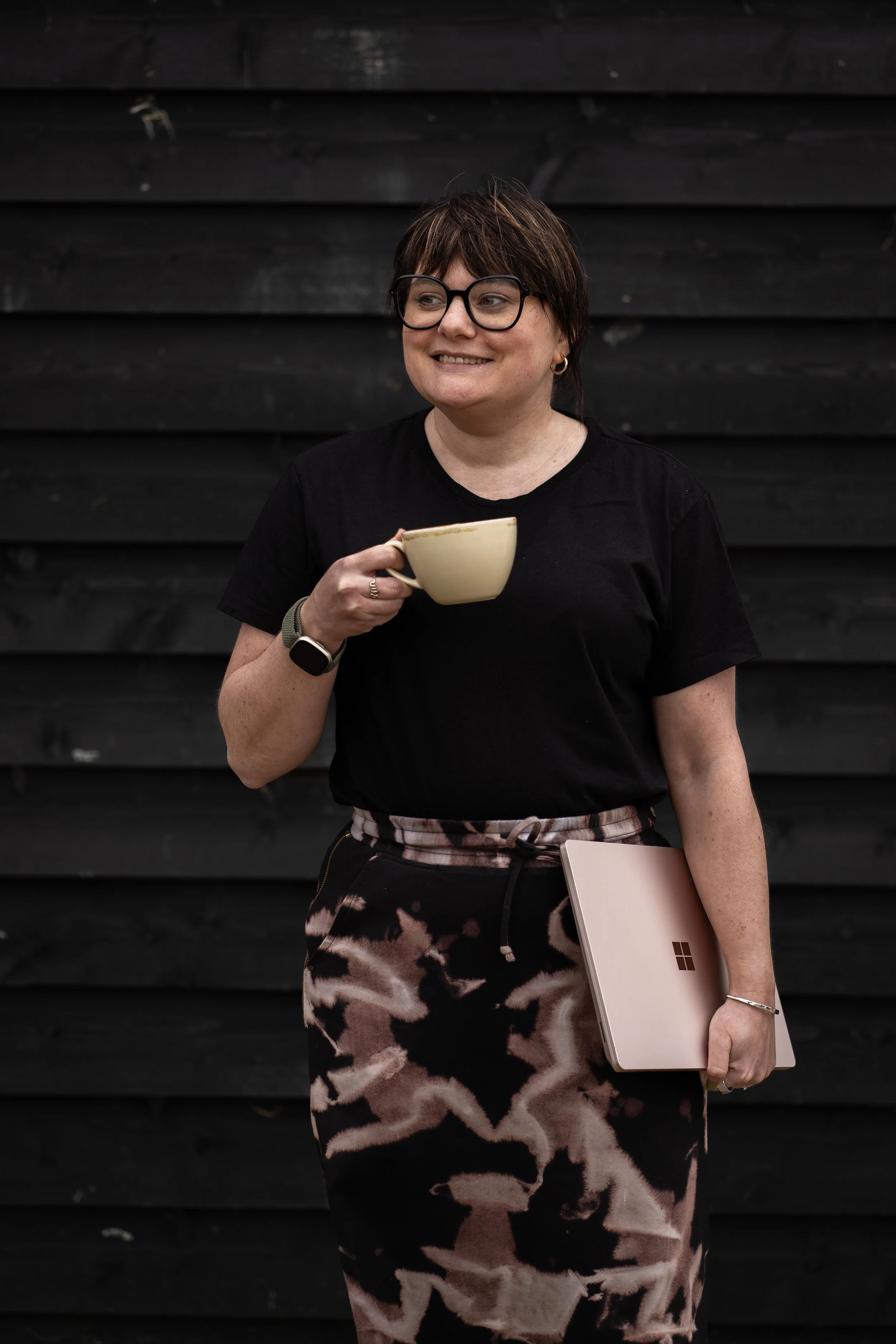 A white woman with short brown hair, wearing glasses, a black t-shirt, and a patterned skirt, holding a cup and a laptop, standing against a black wooden wall ready to start the day as a Virtual Assistant