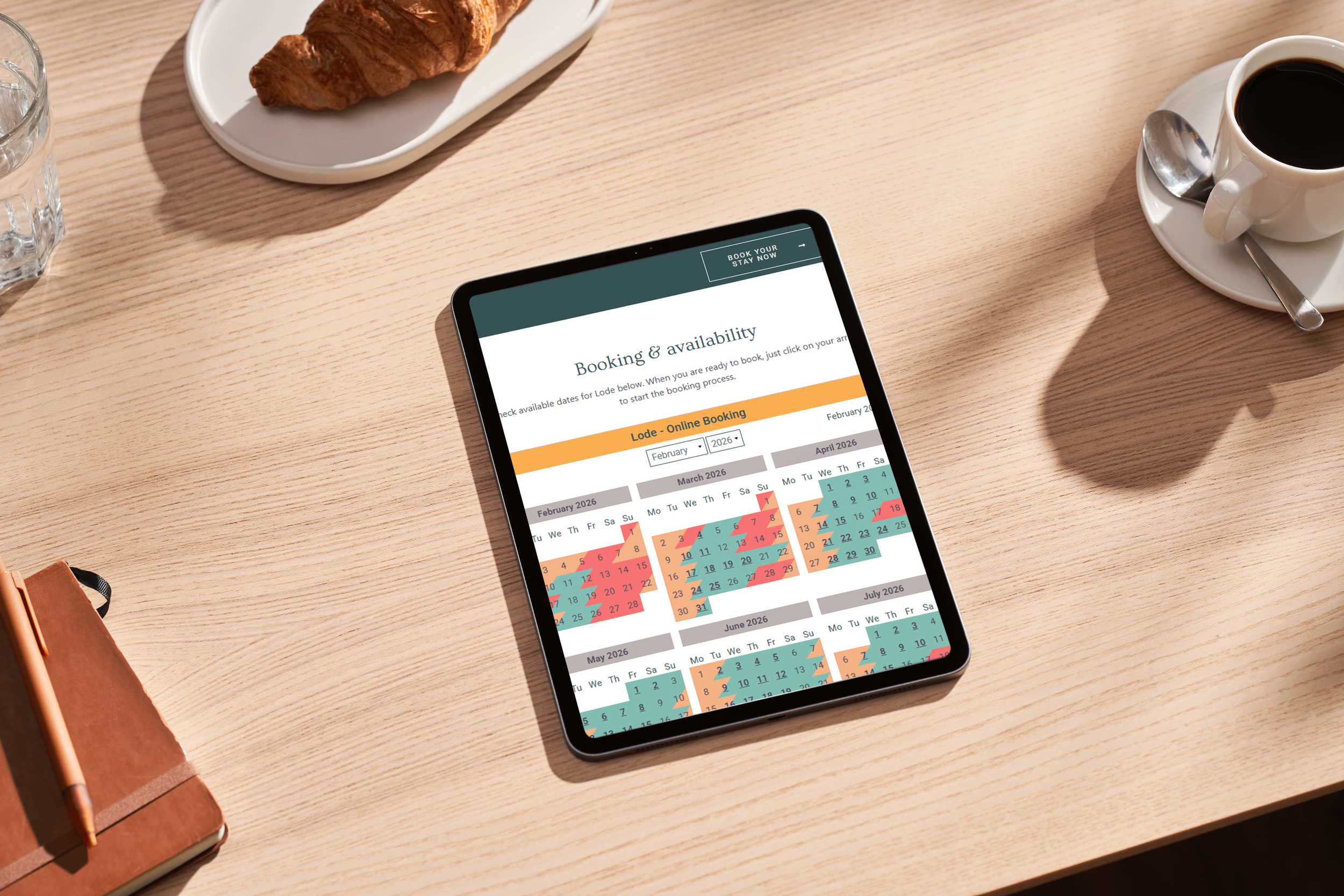 Tablet displaying a booking calendar for lodge reservations with available dates highlighted, on a wooden table along with a cup of coffee, a croissant on a plate, a glass of water, and a closed notebook with a pen.