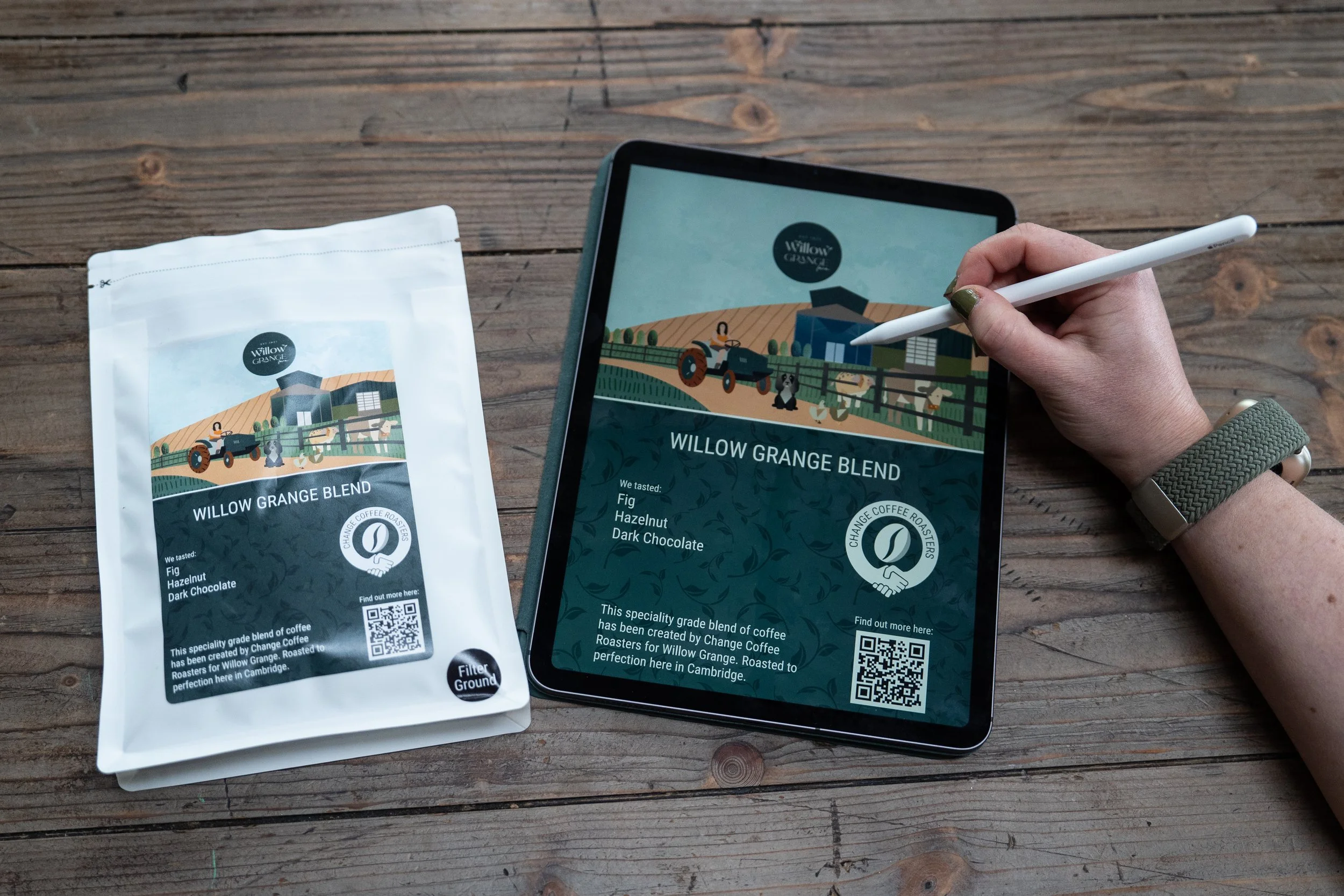 An electronic tablet displaying a coffee product label for Willow Grange Blend, featuring a farm scene with a tractor, animals, and a barn. Next to the tablet is a white coffee bag with the same label, placed on a wooden surface. A person's hand with a smartwatch is holding a stylus and interacting with the tablet.