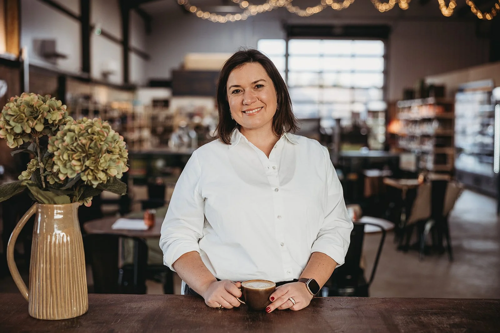 A woman with dark brown hair, wearing a white button-up shirt, holding a coffee mug, standing in a cozy cafe with wooden shelves and tables, a vase with hydrangeas on the counter, and warm ambient lighting.