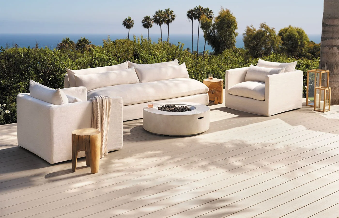 Outdoor patio with white furniture, a sofa and armchair, a round fire pit with black stones, wooden side tables, and scenic view of trees and ocean in the background.
