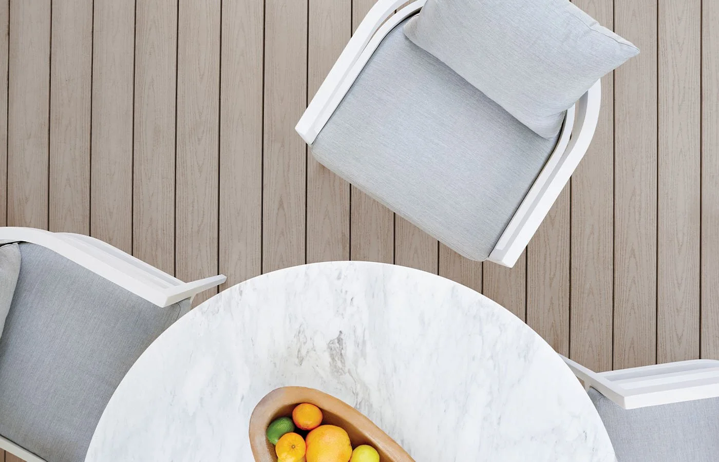 A top-down view of a white marble table with a wooden bowl of assorted citrus fruits and three light gray chairs with cushions on a wooden deck.