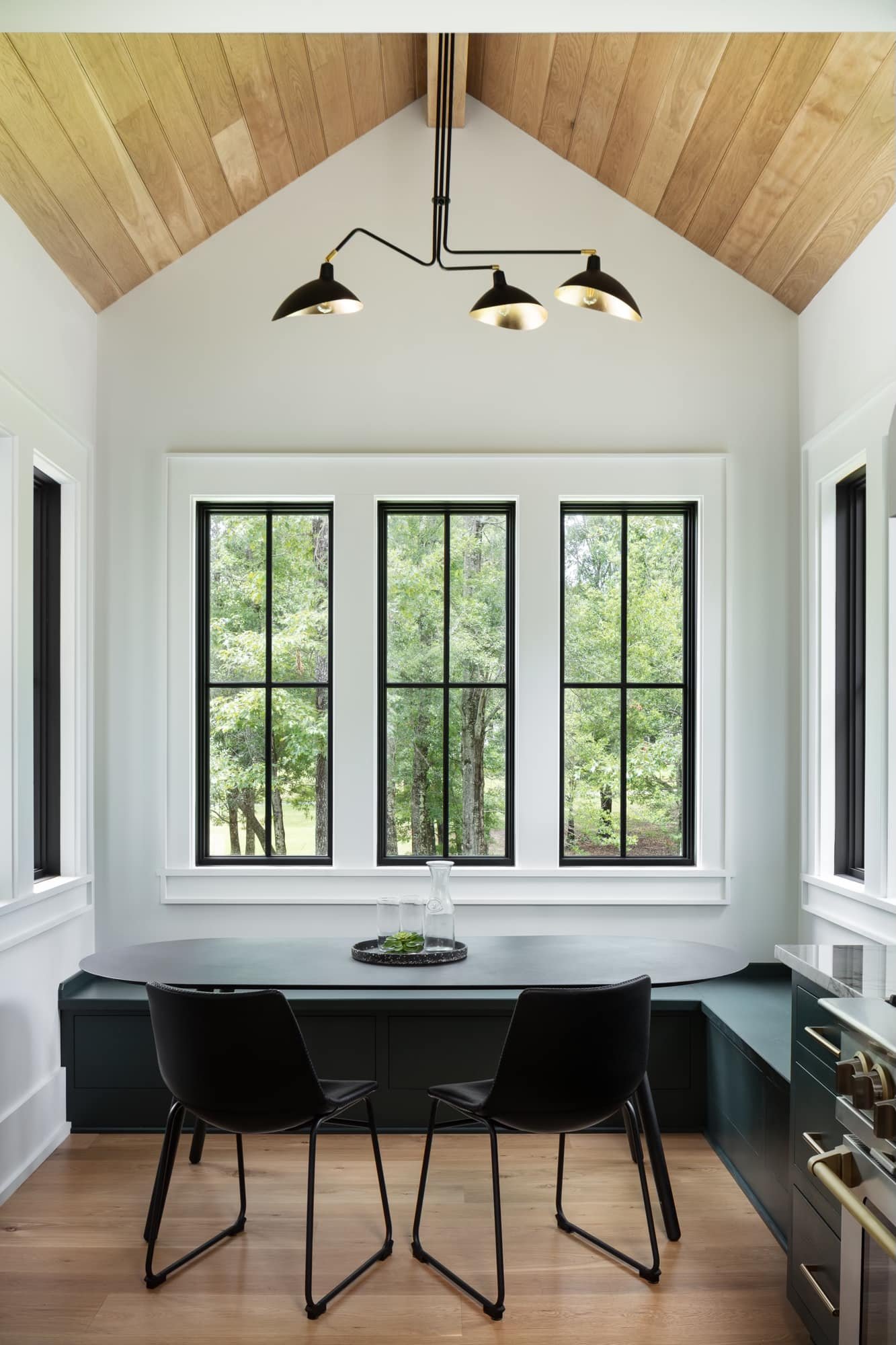 Modern kitchen with large windows, black chairs, a black table, and a black overhead light fixture, with wooden ceiling and light hardwood flooring.
