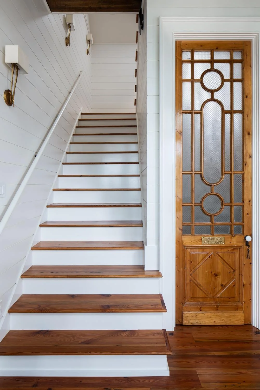 Interior view of a staircase with white painted walls, wooden steps, and brass wall sconces, next to a wooden and glass door with decorative design.
