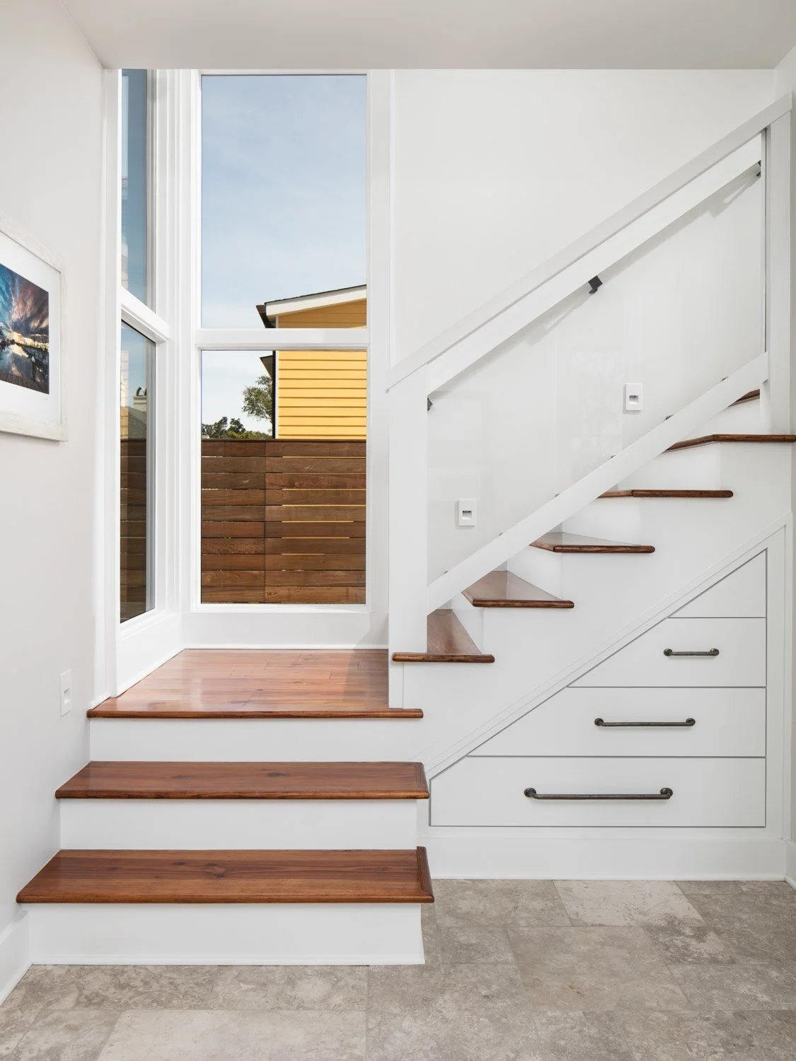 Interior view of a staircase with wooden steps, white risers, and pull-out drawers underneath, leading up to a door with a large window showing a backyard with a wooden fence, a yellow house, and a blue sky.