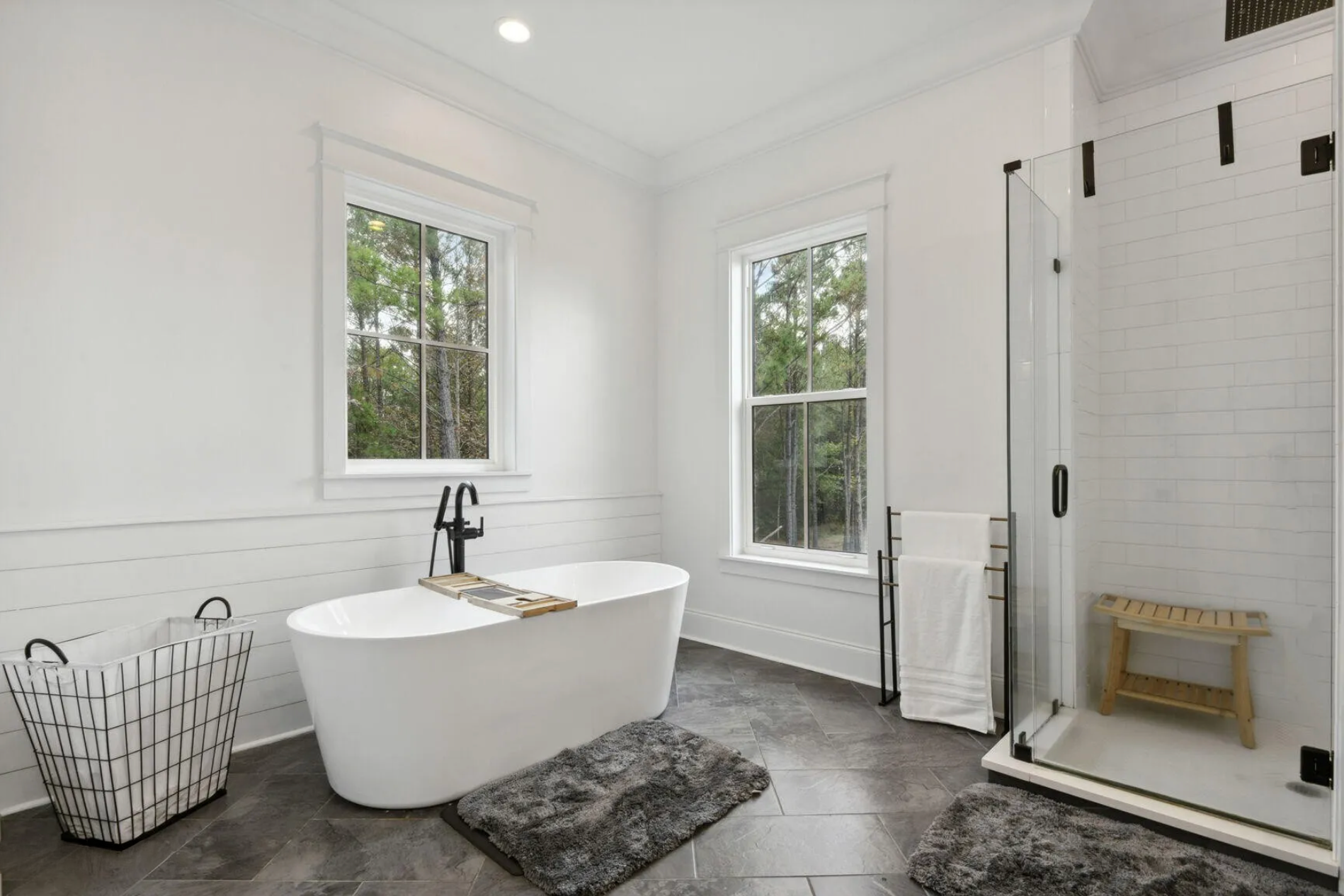Modern bathroom with white walls, a freestanding bathtub, double windows, a glass-enclosed shower, and gray tile flooring.