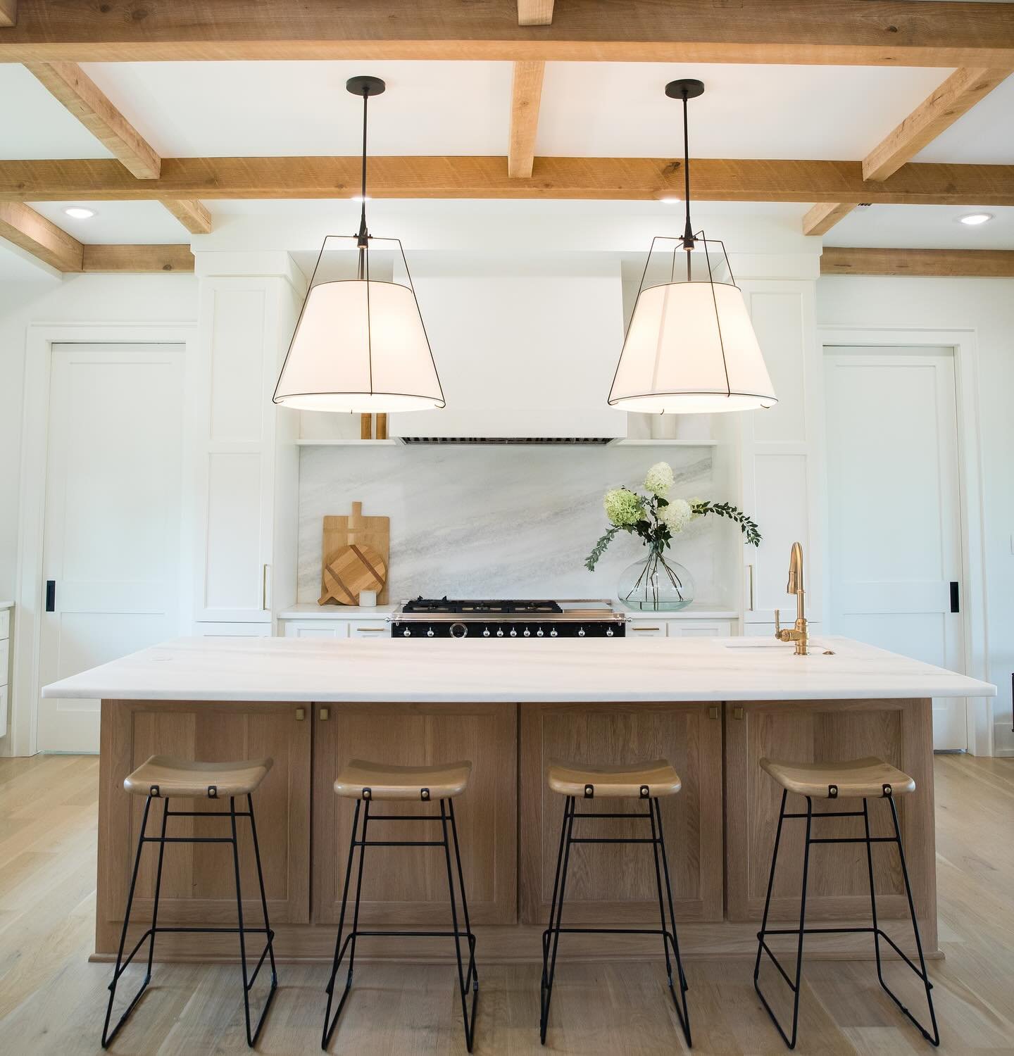 Modern kitchen with a white island, three bar stools, two hanging pendant lights, a gas stove, a white marble backsplash, a glass vase with white flowers, and wooden accents.