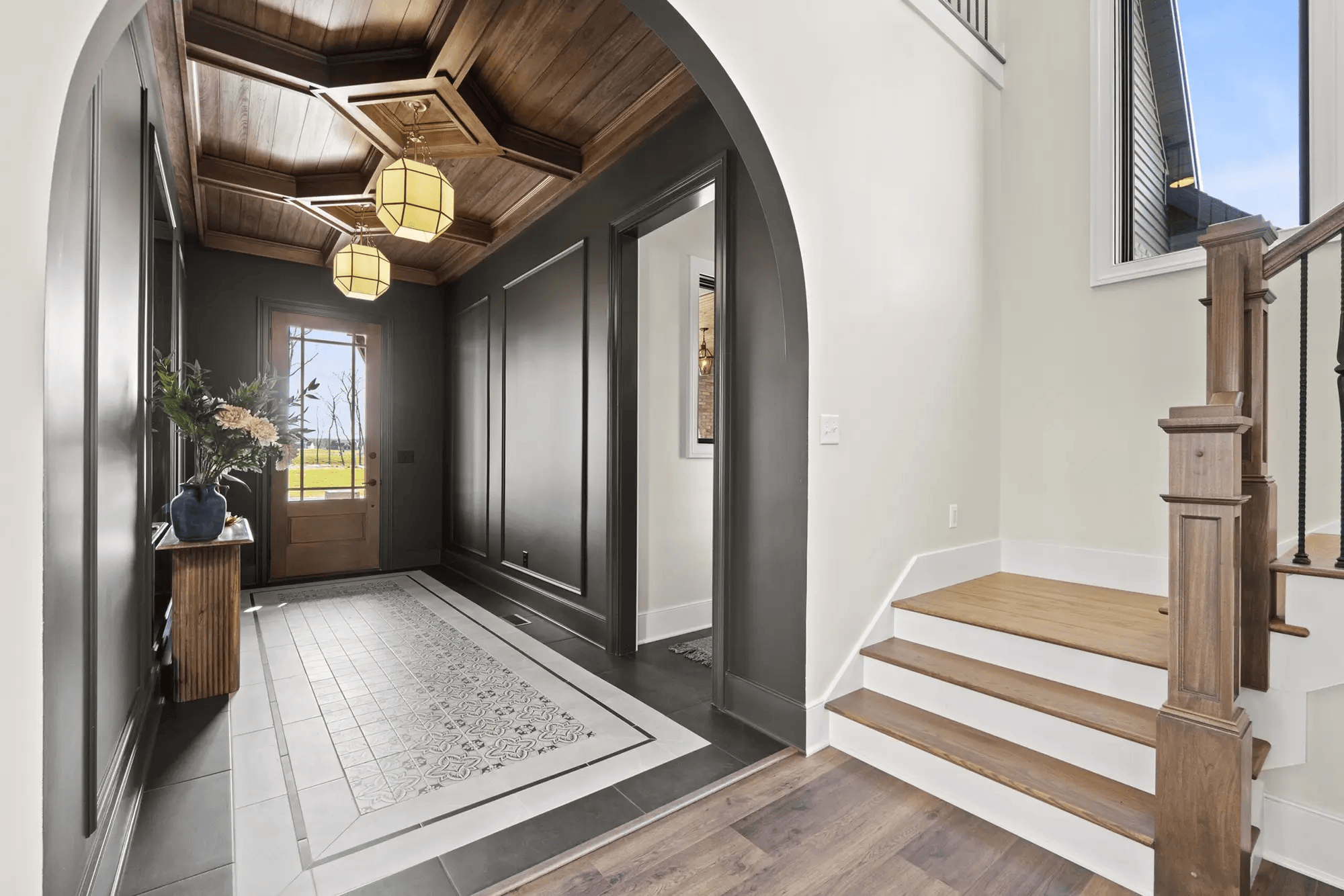 Entry hallway with a wooden and gray interior, decorated with geometric hanging lights, an arched doorway, a small wooden table with a vase of flowers, and a window showing a scenic outdoor view.