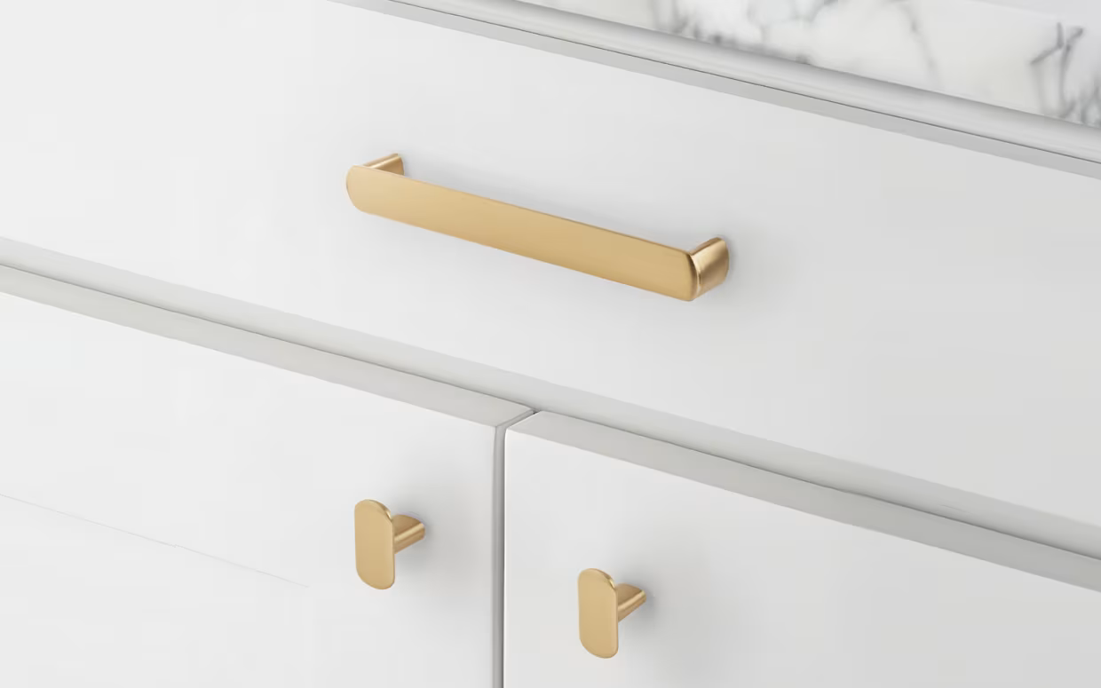 Close-up of a white cabinet with gold handles, featuring a horizontal handle on a drawer and two smaller vertical handles on cabinet doors.