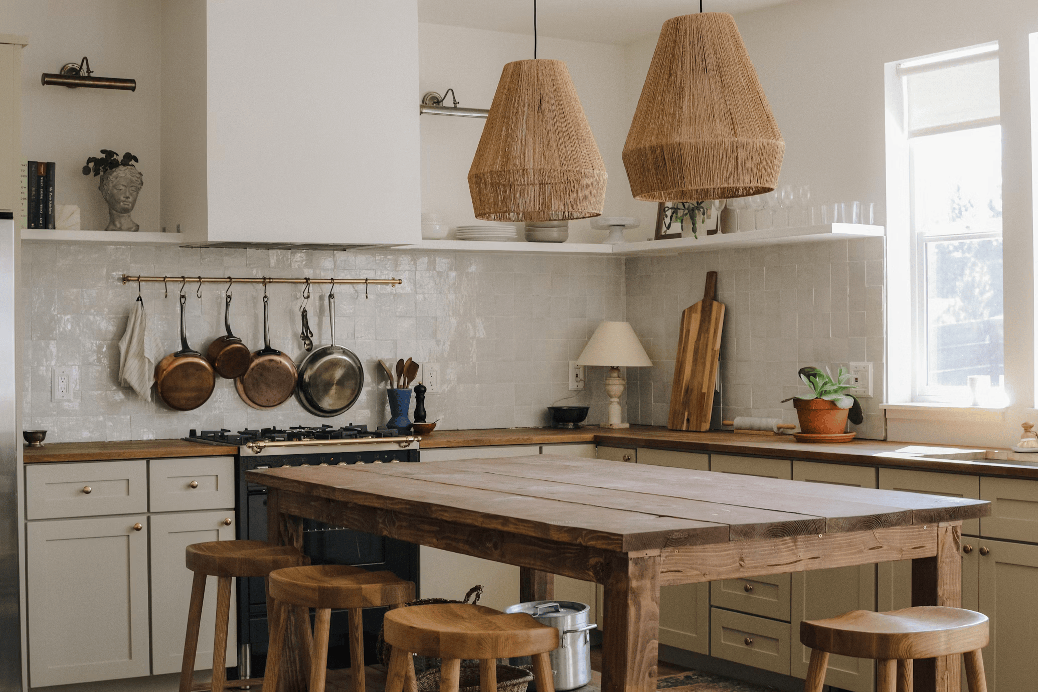A cozy kitchen with cream-colored cabinetry, a wooden dining table, and four matching wooden stools, decorated with hanging copper pots, a cutting board, a potted plant, and pendant lights with woven shades.