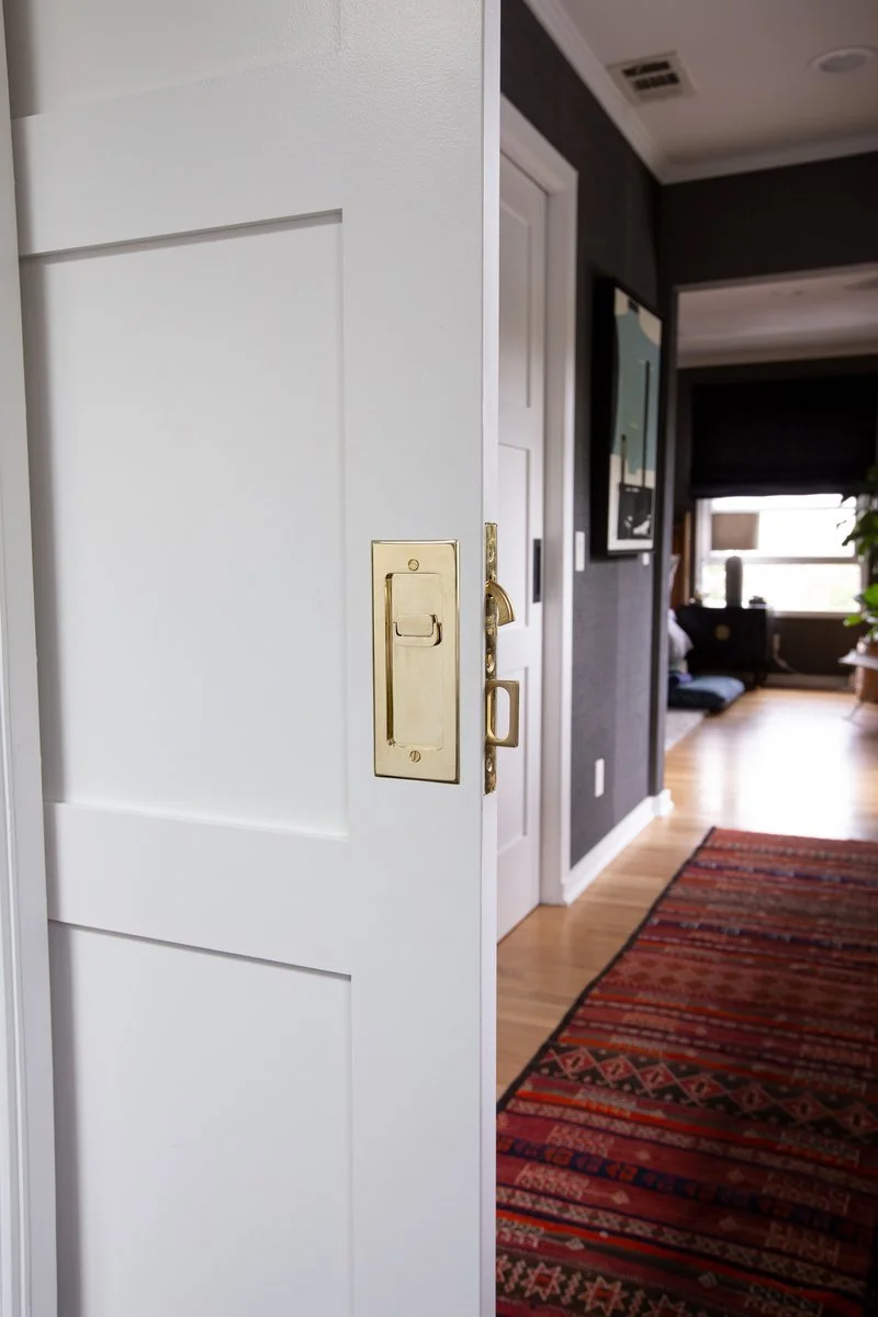 Partially open white door with brass locks in a home interior, revealing a hallway with wooden flooring, a patterned rug, a wall-mounted painting, and a living room with a window and furniture in the background.