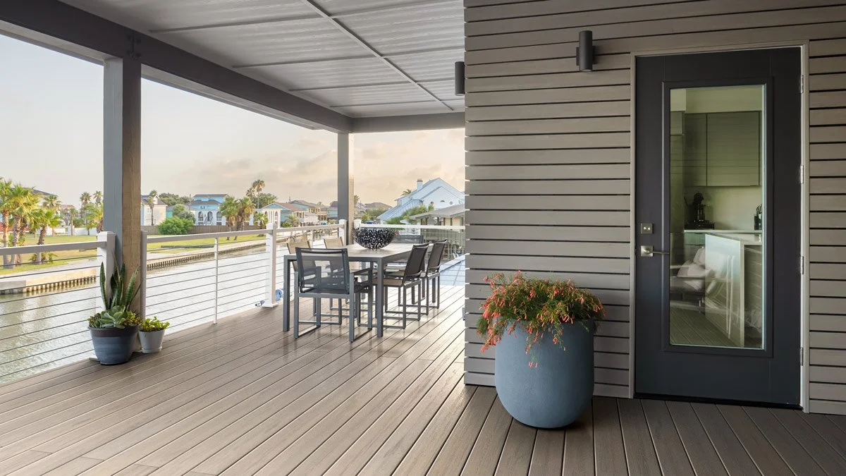 An outdoor balcony with wooden flooring, potted plants, a dining table with chairs, overlooking a waterway and residential neighborhood with palm trees.