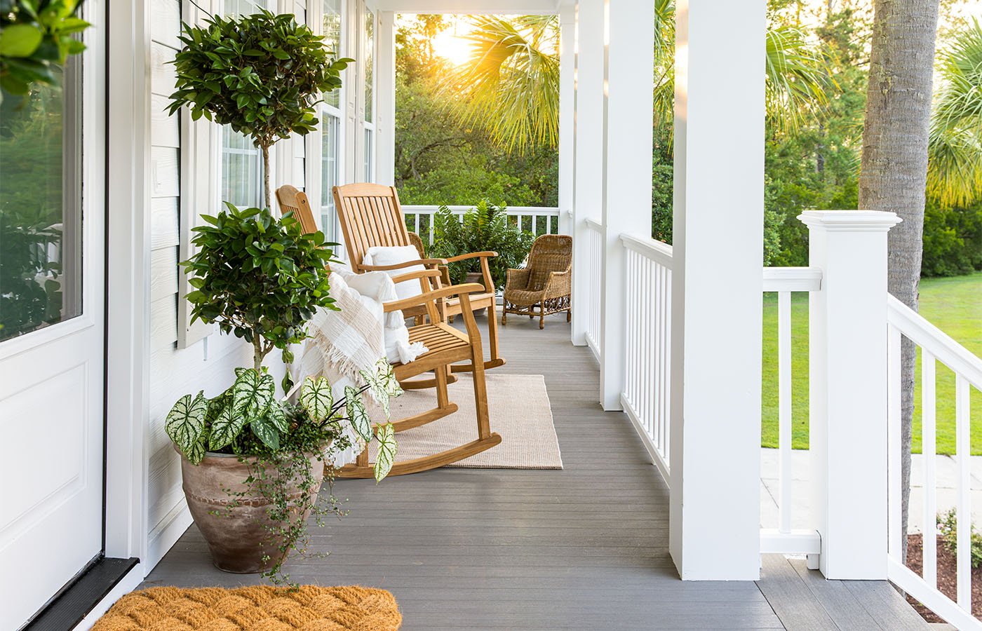 Front porch with wooden rocking chairs, potted plants, and a view of green trees and lawn in the background during sunset.