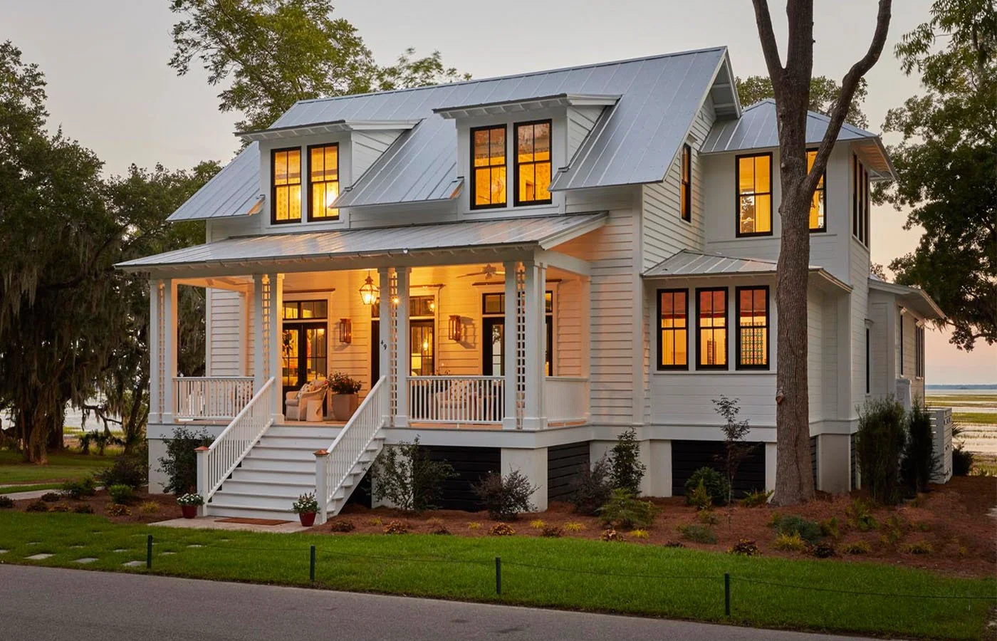 White two-story house with a metal roof and front porch, illuminated interior lights, surrounded by landscaping and tall trees