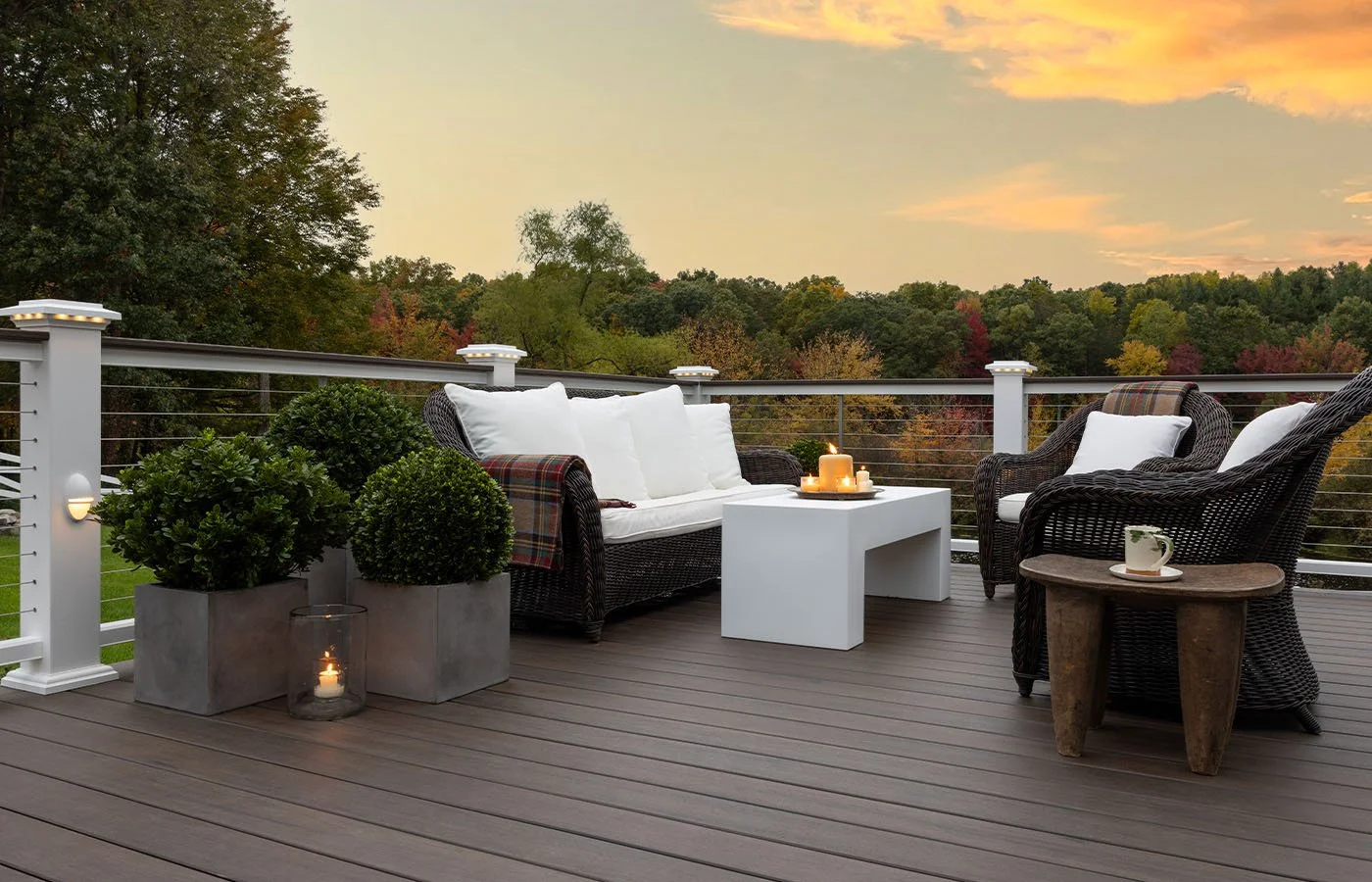 Outdoor deck with wicker furniture, including two armchairs and a sofa with cushions, a white table with candles, potted plants, and a scenic view of trees with fall foliage at sunset.