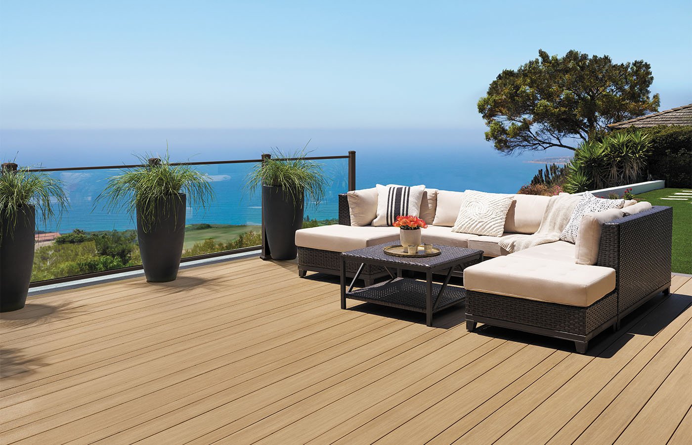 Modern outdoor patio with beige sectional sofa, coffee table, and potted plants, overlooking blue ocean and sky.