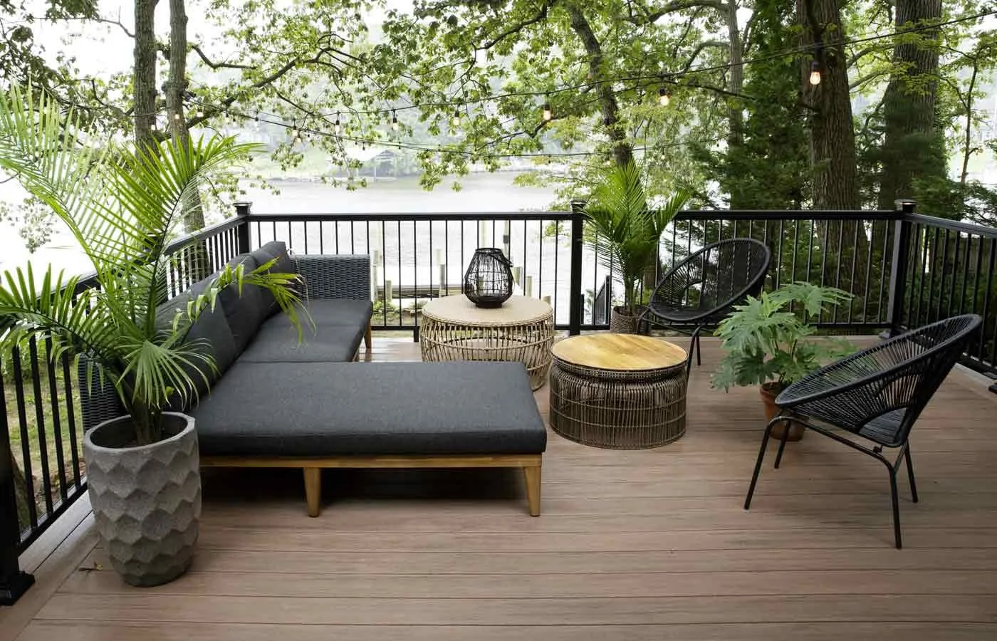 Outdoor balcony with black railing, wooden floor, black sofa, two round woven side tables, black chairs with rounded backs, potted green plants, string lights, overlooking trees and water.
