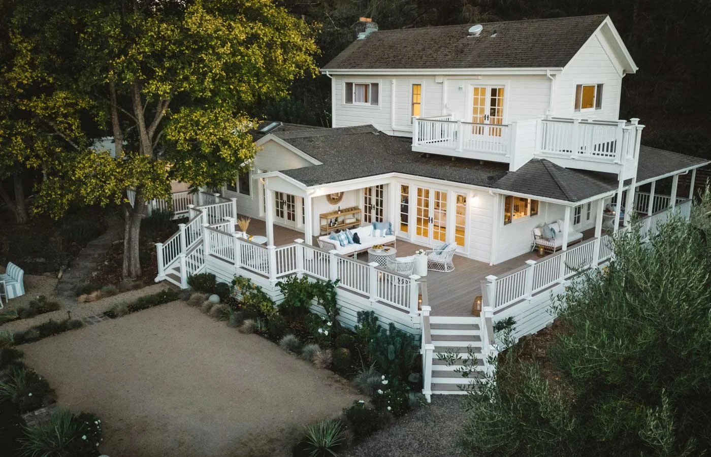 A large white house with a wraparound deck, multiple patio furniture sets, and several large trees around the yard, viewed from above during dusk.