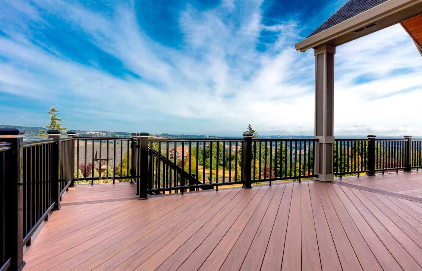 View from a wooden deck with black railing, overlooking a suburban area with houses, trees, and a cloudy blue sky.