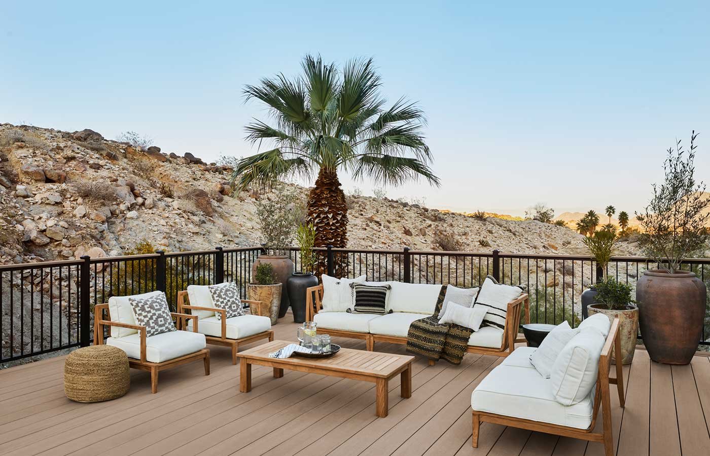 Outdoor patio with wooden furniture, white cushions, and large potted plants, overlooking rocky desert landscape with palm trees in the background.