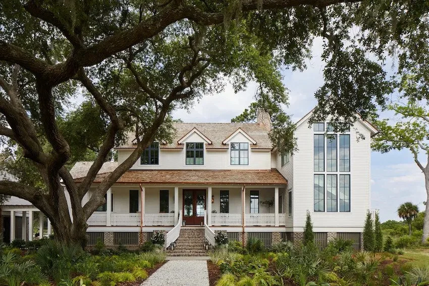 Large white house with multiple windows and a front porch, surrounded by leafy trees and a landscaped garden.