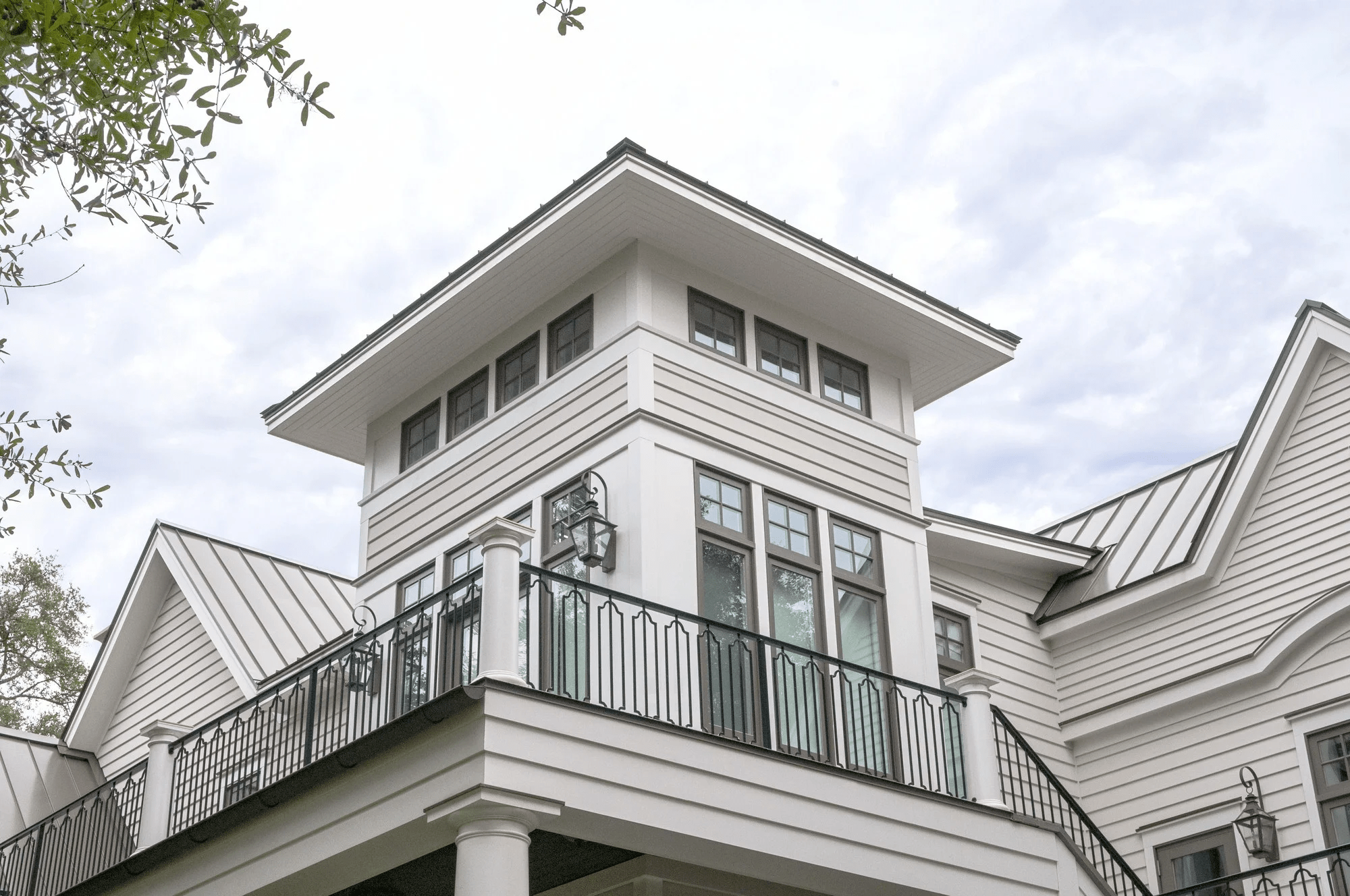 A white modern house with multiple rooflines and a balcony with black railing, multiple windows, and outdoor lanterns under a cloudy sky.