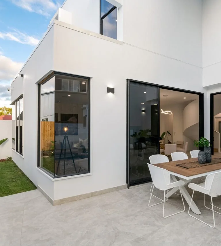 Modern white house with large windows and patio, outdoor dining table with chairs, and interior visible through sliding glass doors.