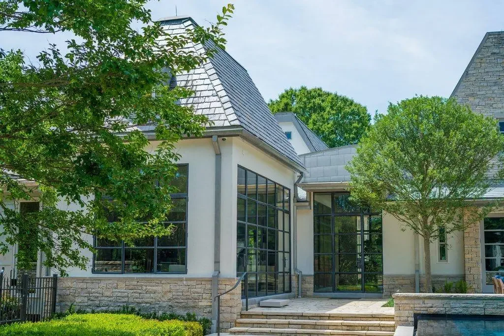 Front view of a modern house with large glass windows, brick and white exterior walls, and surrounded by trees and greenery.