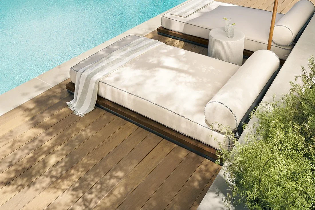 Two white cushioned lounge chairs by a swimming pool with a small white side table between them, holding a glass with drink. There is a striped towel on one lounge chair, and a wooden pool umbrella. Shadows of trees are cast on the chairs and the wooden deck, with some greenery on the right.