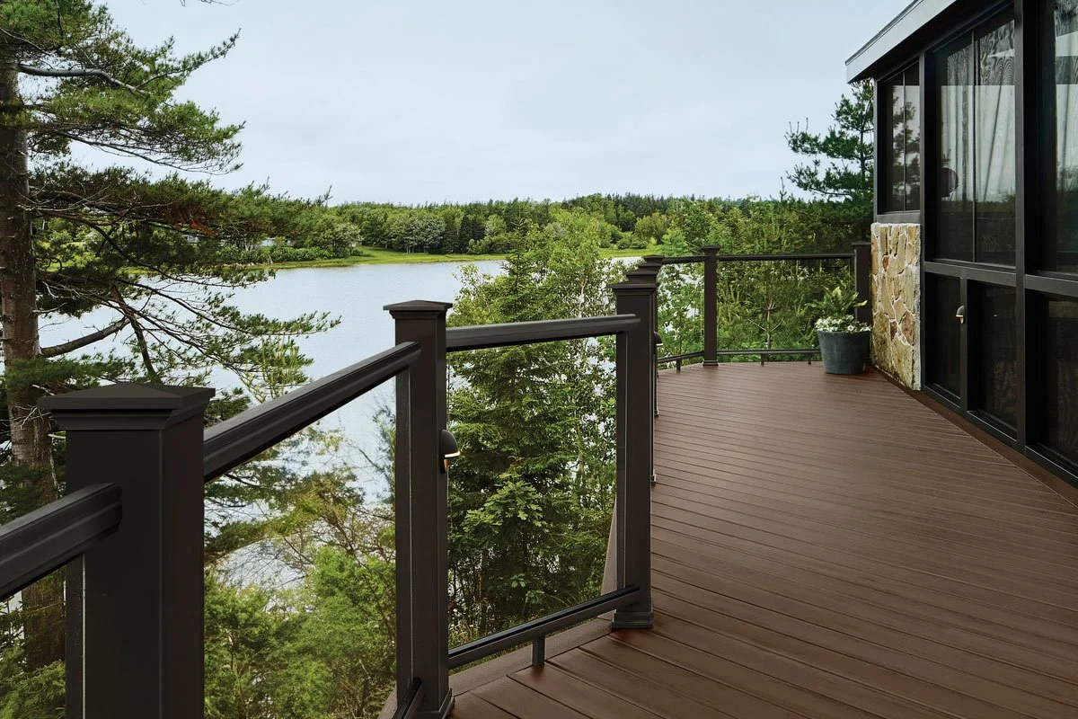 View of a wooden deck overlooking a lake surrounded by trees, with a black metal railing and a potted plant near the house.