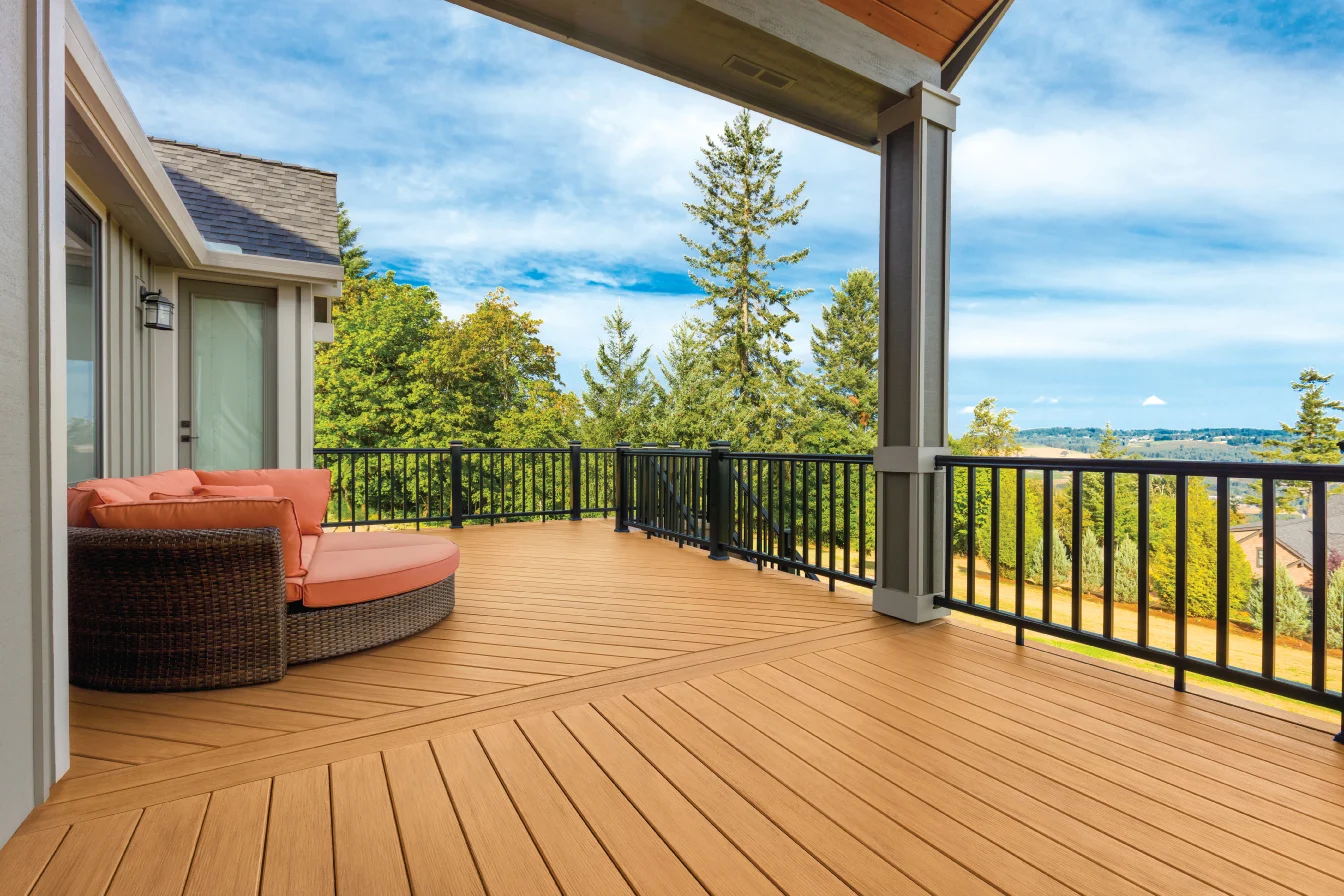 Back porch with wooden deck, black railing, peach outdoor sofa, and view of green trees and a blue sky with some clouds.