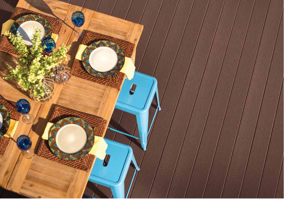 Dining table set for four with plates, bowls, glasses, yellow napkins, and a floral centerpiece, on a wooden deck with two blue stools.